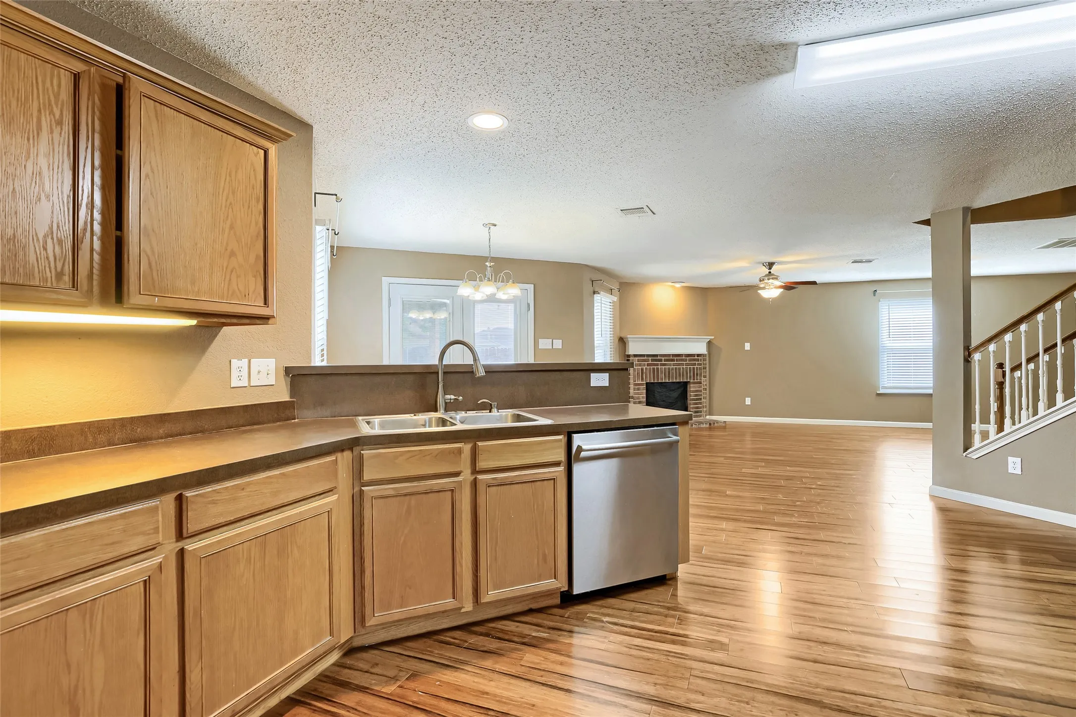 Kitchen featuring wood cabinetry, stainless steel dishwasher, double-basin sink, wood-finish flooring, and brick fireplace