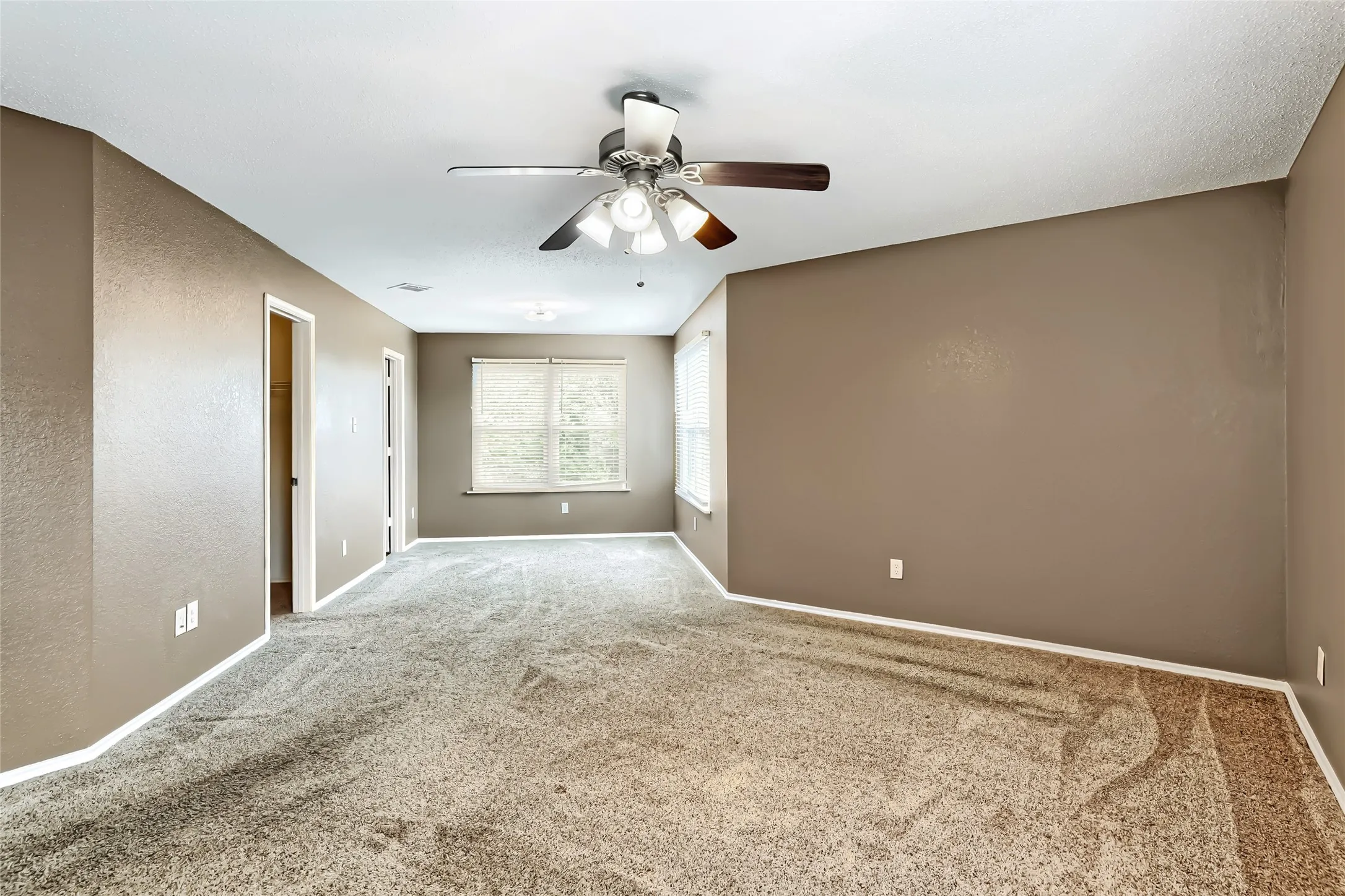 Neutral-toned interior space featuring wall-to-wall carpeting, dual windows with blinds, and a multi-blade ceiling fan with integrated lighting