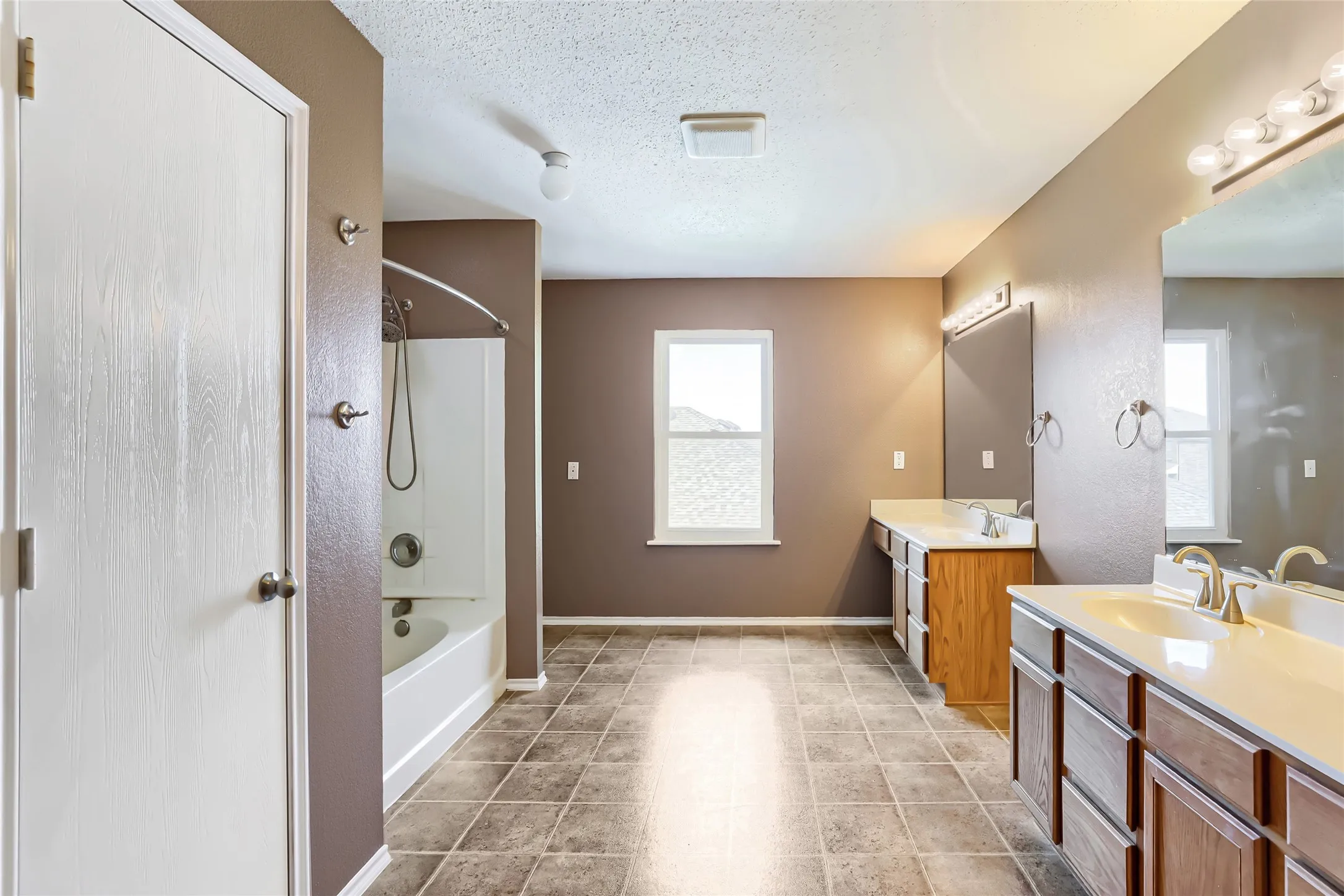 Bathroom featuring tile flooring, dual-basin vanity with wood-finish cabinetry, integrated mirrors with vanity lighting, and a shower-tub combination