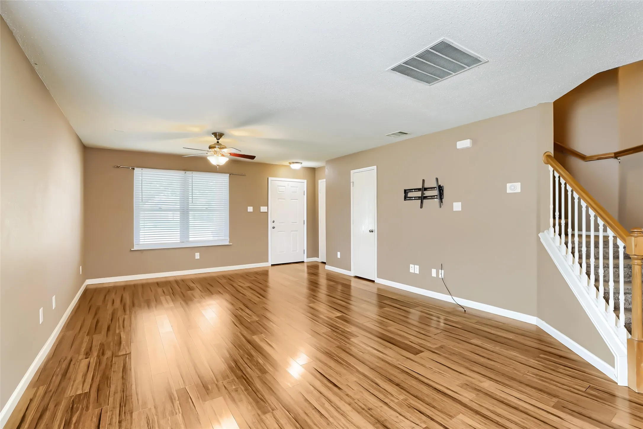 Wood-finish flooring, ceiling fan with integrated lighting, window with blinds, and staircase featuring white balusters with a wood-finish handrail