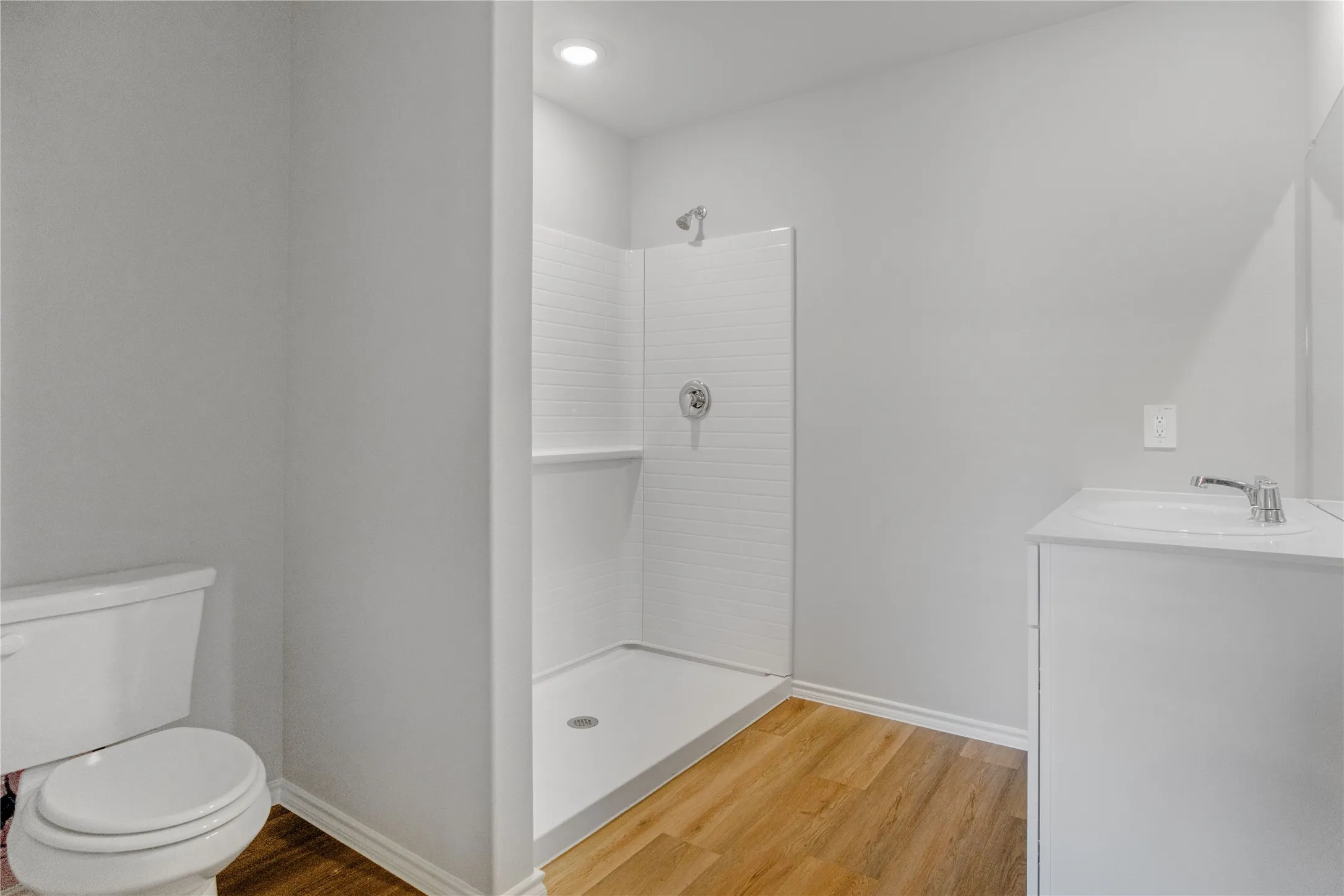 Bathroom featuring a white vanity with a integrated sink, a walk-in shower with tiled surround, wood-finish flooring, and recessed lighting