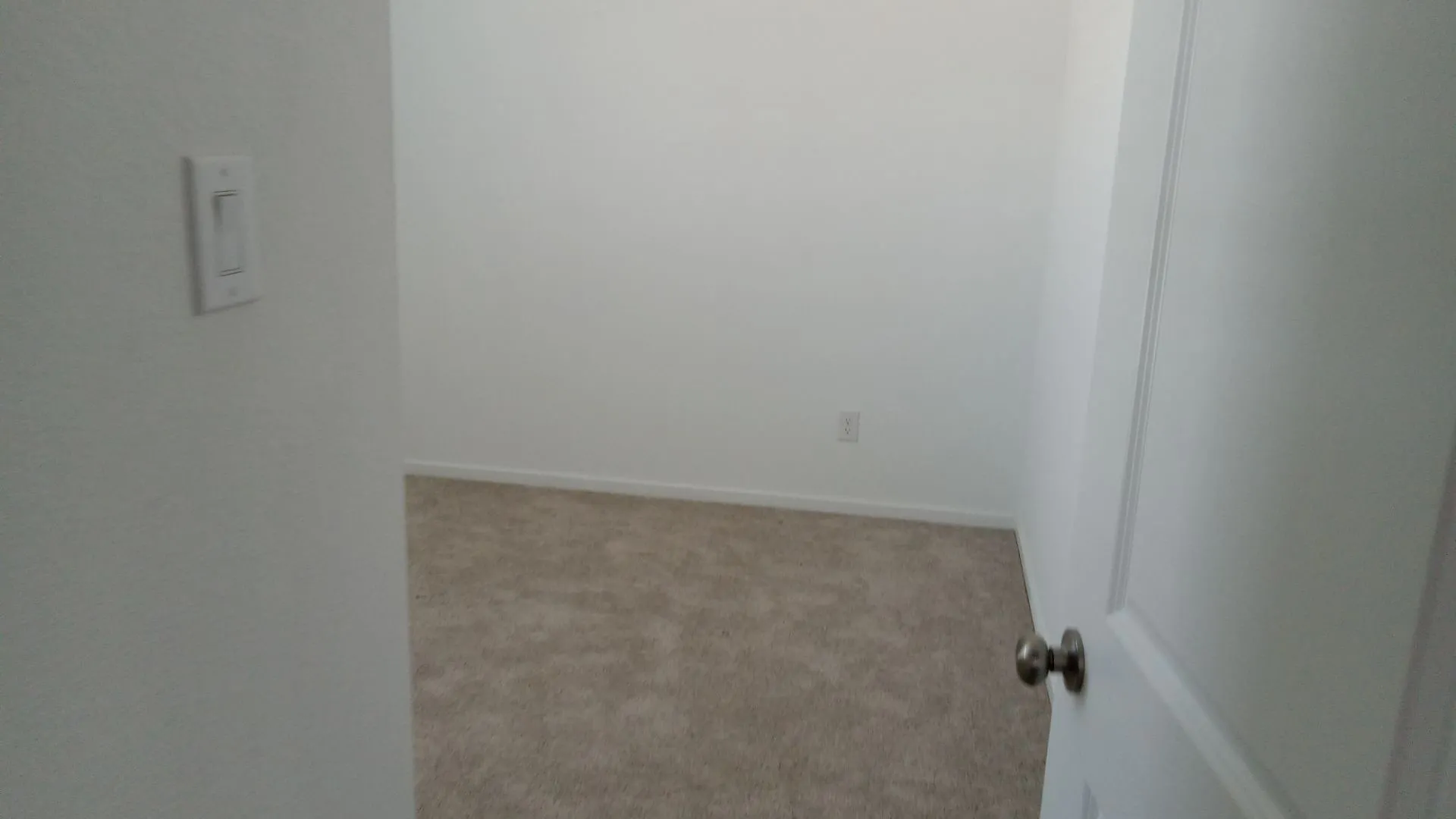 Interior space featuring neutral wall paint, wall-to-wall carpeting, a multi-panel door with a metallic knob, and integrated electrical switch and outlet​​‌​​​​‌​​‌‌​‌‌​​​‌‌​‌​‌​‌​​​‌​​ hardware