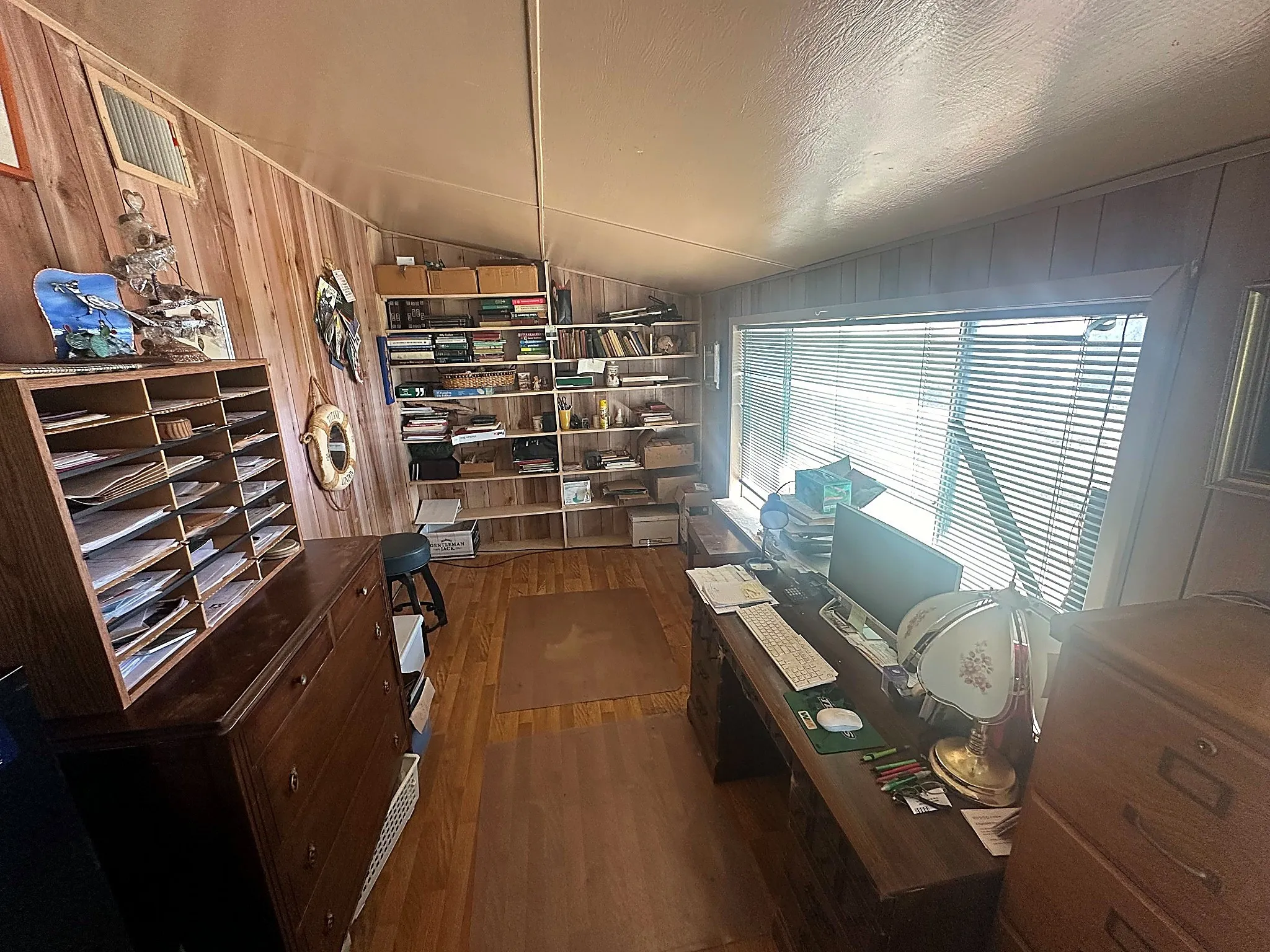 Wood-paneled walls, built-in shelving units, wide horizontal window with blinds, and wood-finish flooring
