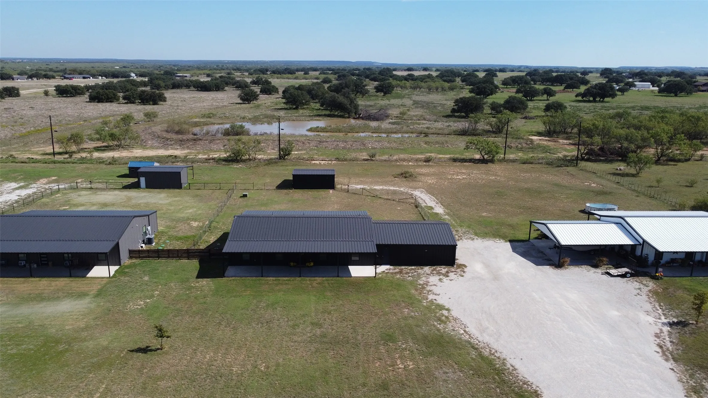 Aerial perspective of expansive acreage featuring multiple metal-roofed structures, fenced enclosures, and a gravel driveway