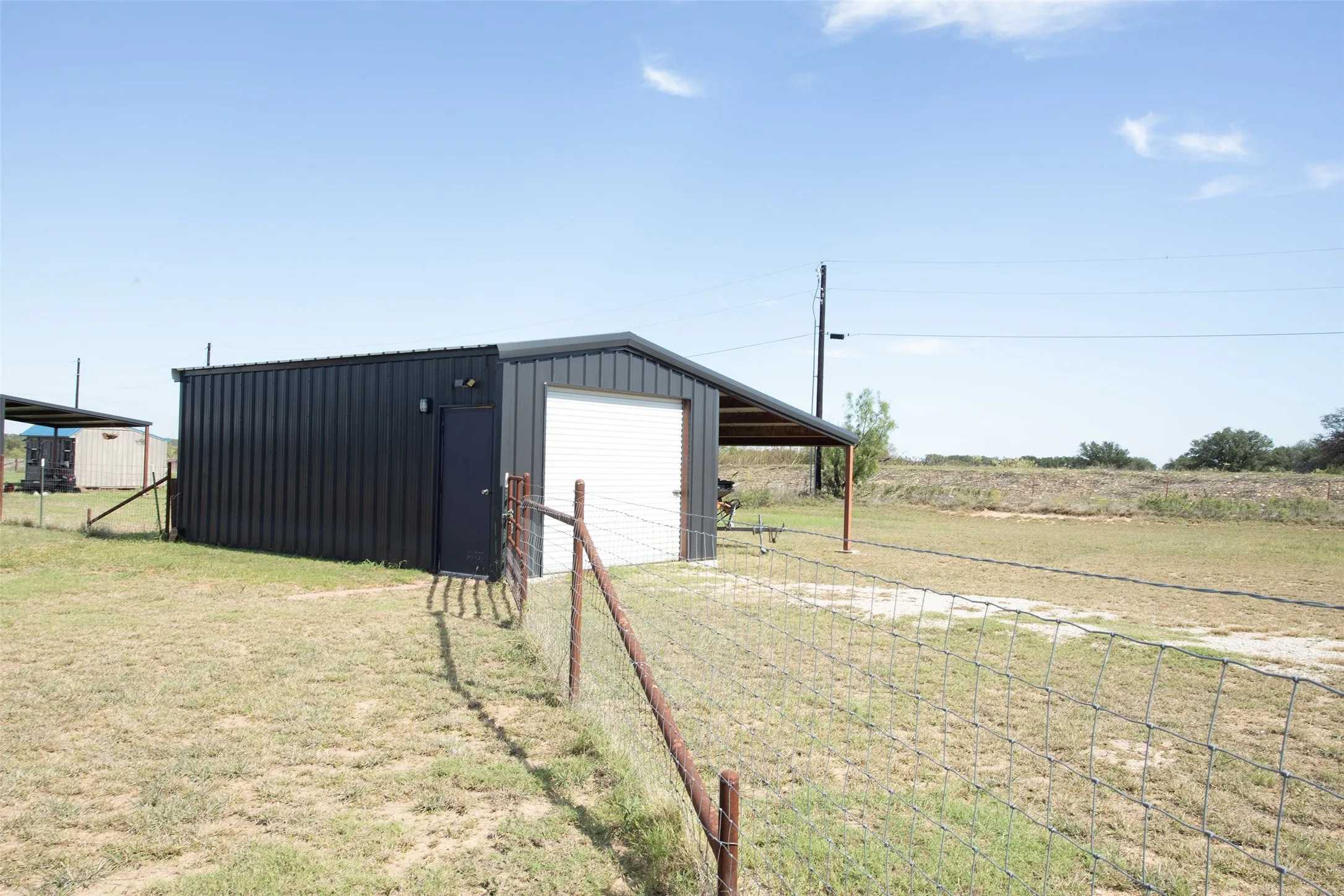 Metal-sided outbuilding featuring a roll-up garage door, an entry door, and an attached covered lean-to on a fenced lot