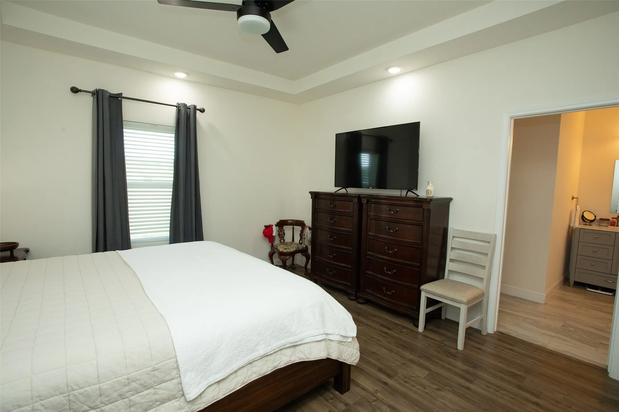 Tray ceiling with recessed lighting, ceiling fan with integrated light fixture, wood-finish flooring, and dual-track window drapery hardware