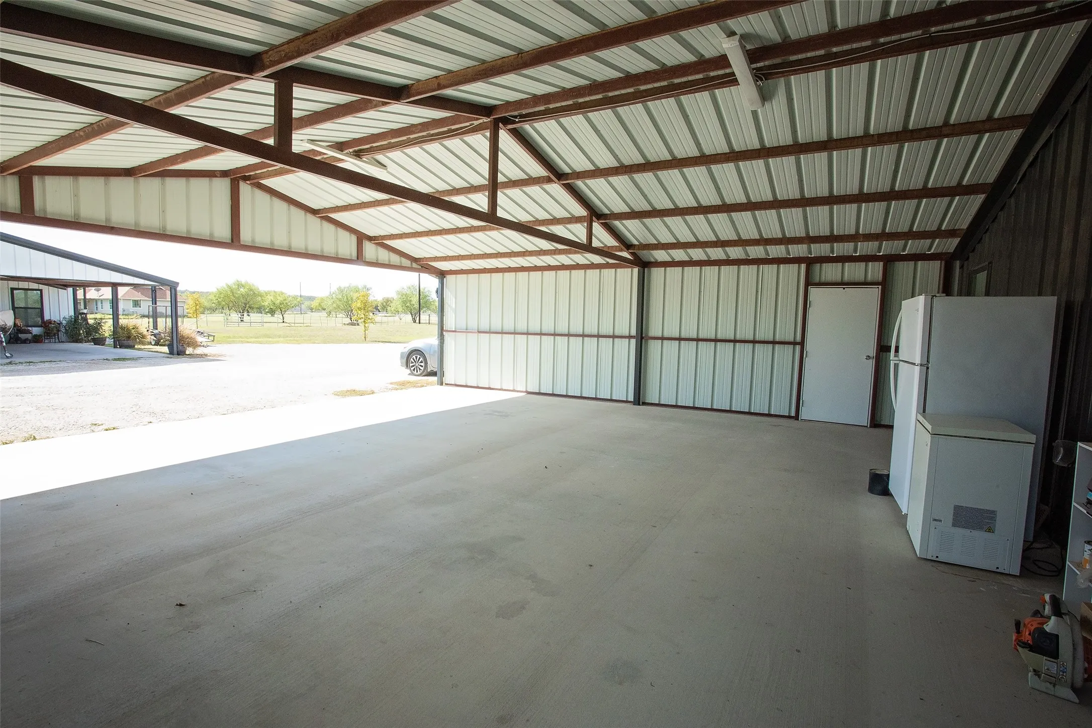 Covered carport featuring a steel frame structure, corrugated metal roofing, concrete flooring, and an enclosed storage door