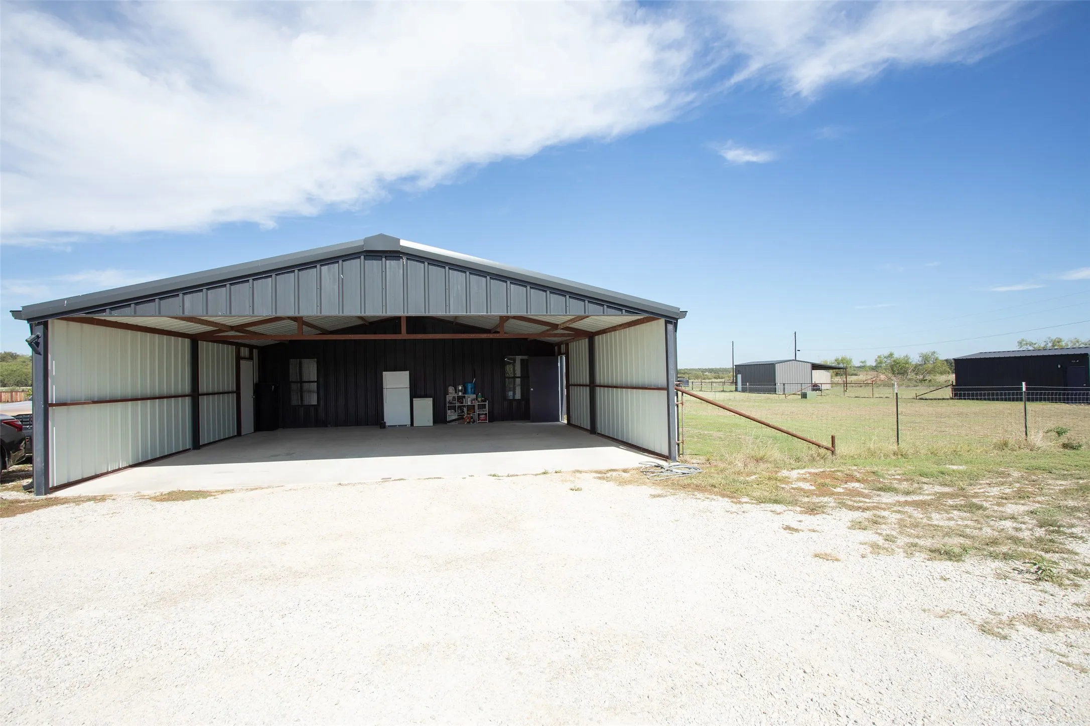 Covered metal-sided workshop featuring a concrete foundation, open-bay design, and expansive gravel parking area