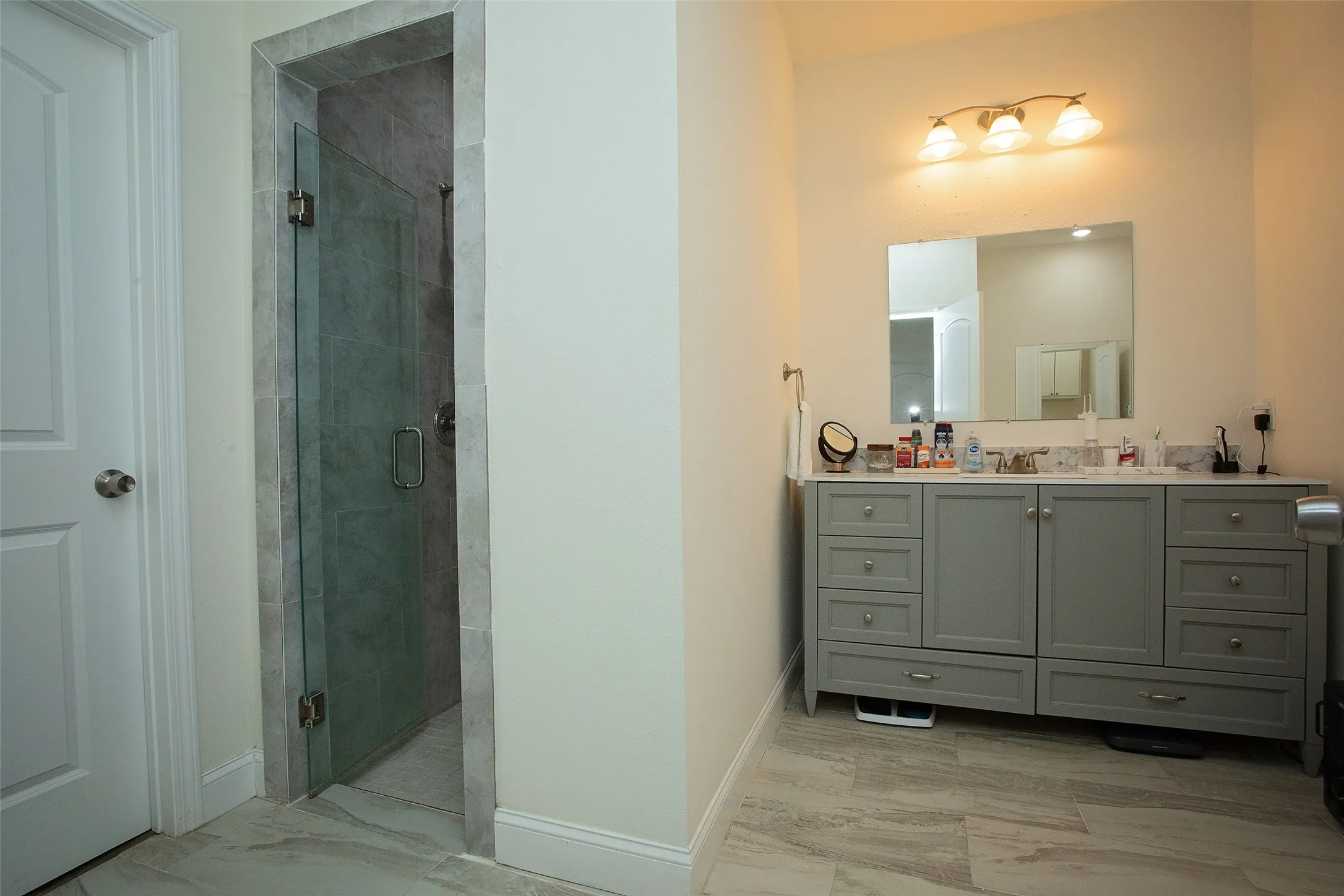 Bathroom featuring a gray vanity with stone-style countertop, tile flooring, frameless glass-enclosed shower with stone tile surround, and wall-mounted vanity lighting