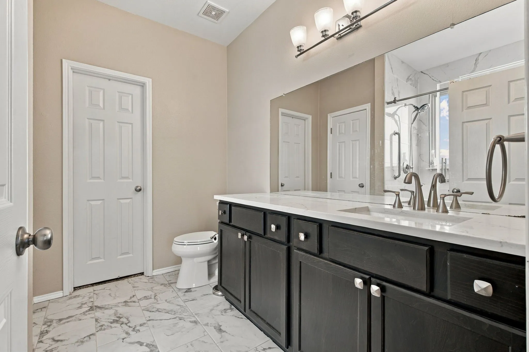 Black vanity cabinet with a light-toned countertop, integrated dual-sink basins, polished metal fixtures, and stone-patterned tile flooring