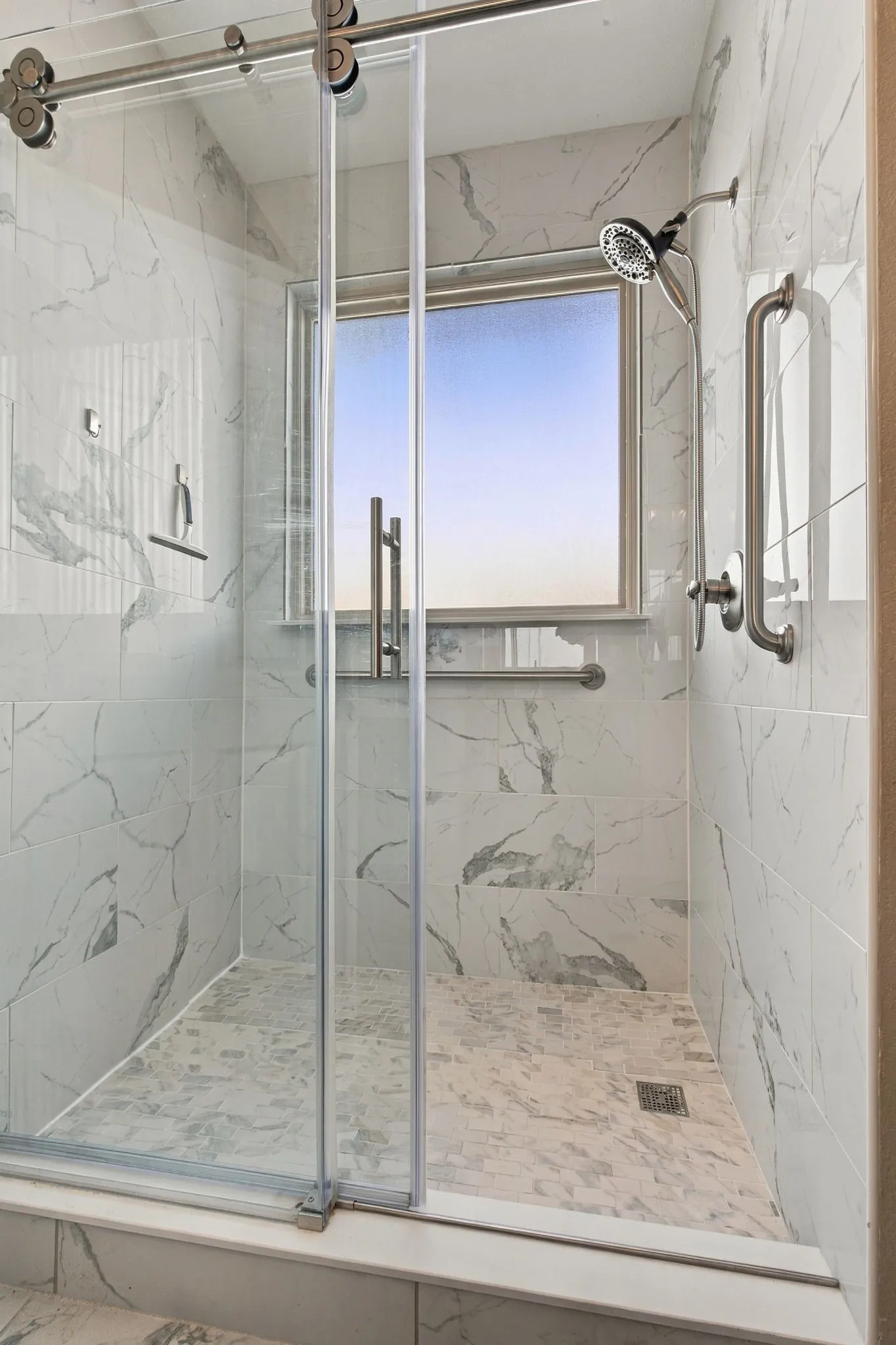 Walk-in shower featuring large-format stone-patterned tile, sliding glass door with stainless steel hardware, integrated grab bars, and a built-in window