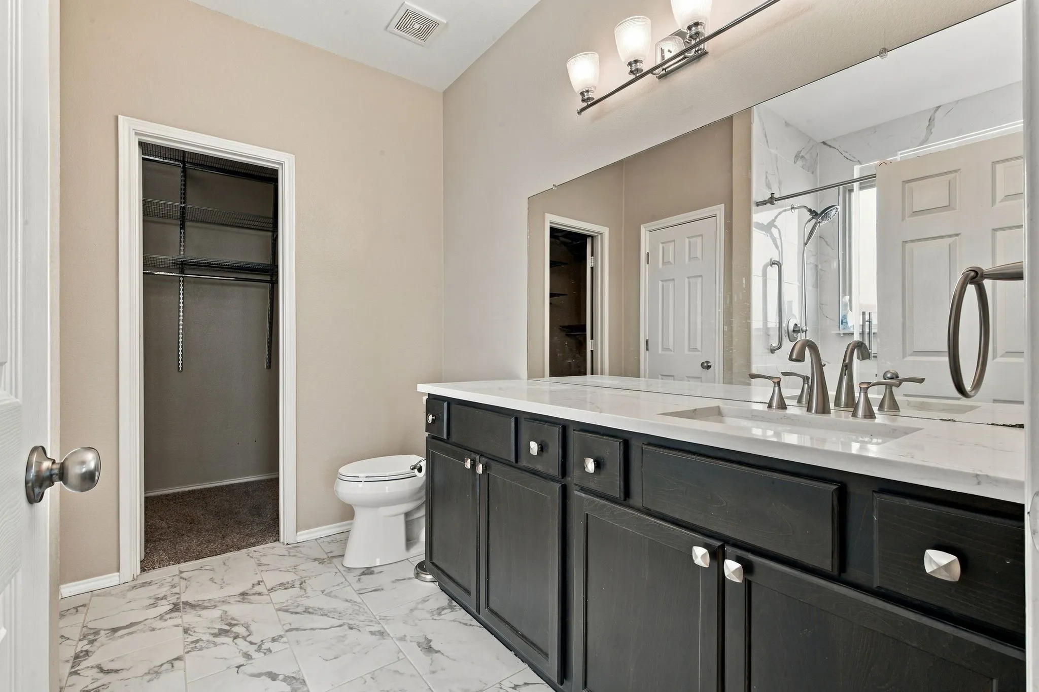 Bathroom featuring dark cabinetry with light stone-style countertops, an integrated vanity mirror, three-light fixture, and stone-finish tile flooring