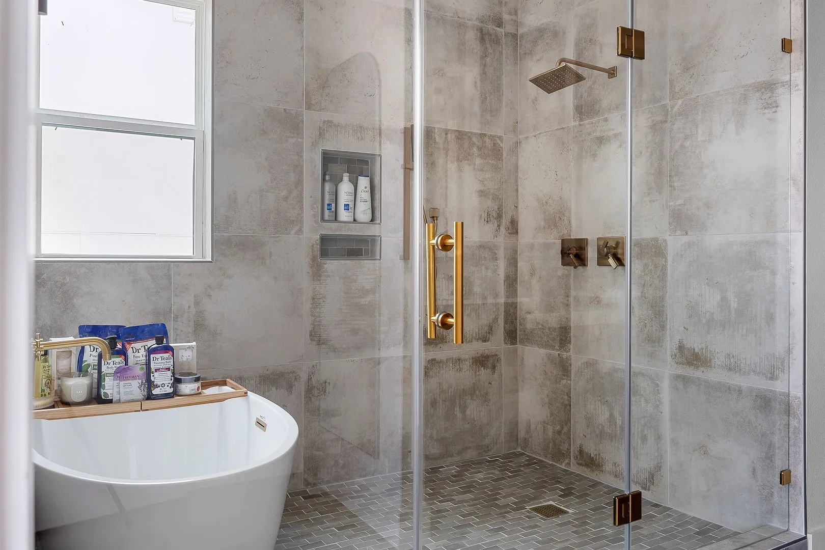 Walk-in shower featuring large-format neutral wall tile, mosaic tile flooring, glass enclosure, brass-finish fixtures, and integrated wall niches