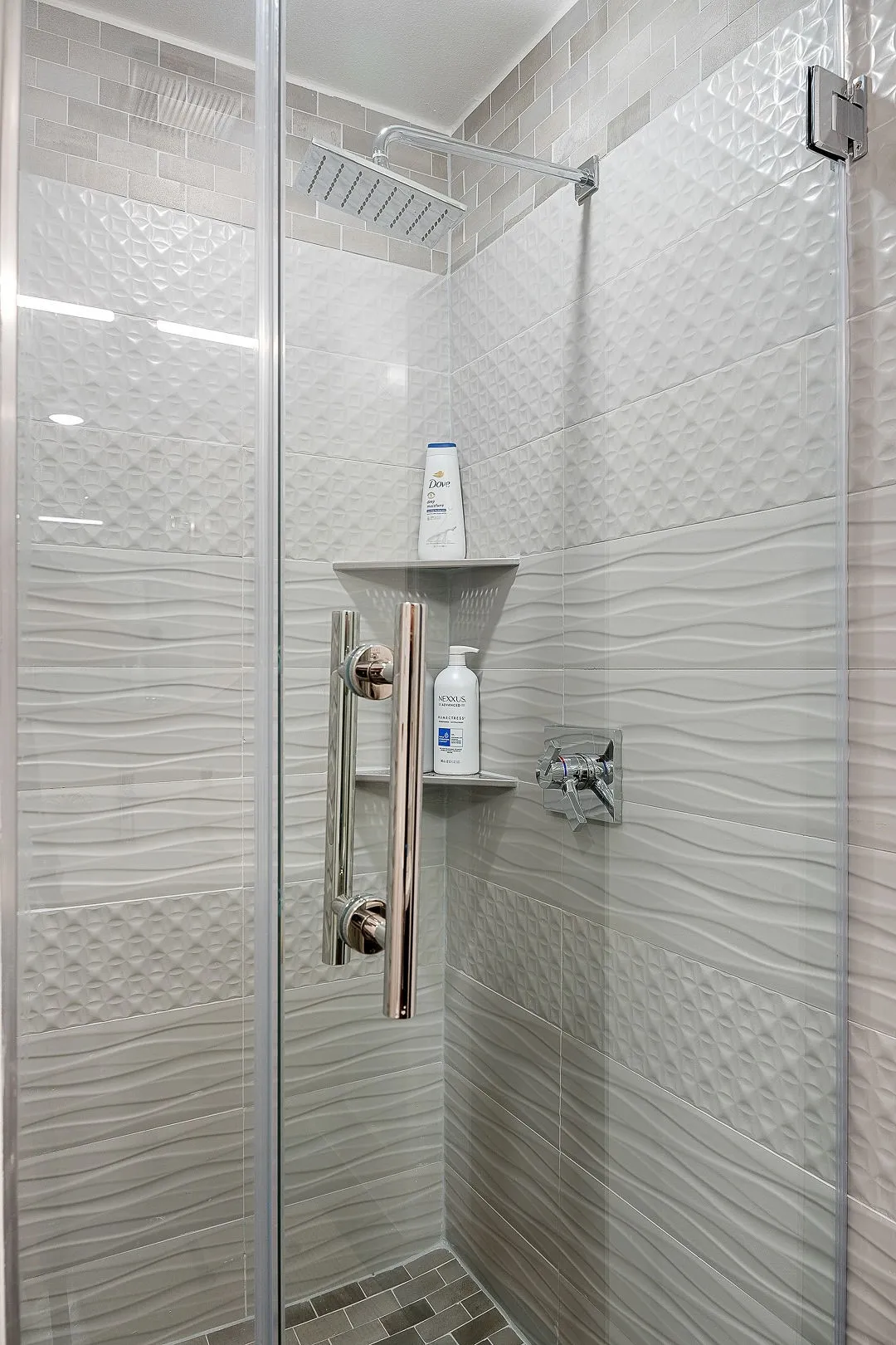 Walk-in shower featuring a frameless glass door, rain-style showerhead, chrome fixture hardware, tiered corner shelving, and multi-textured tile wall detailing