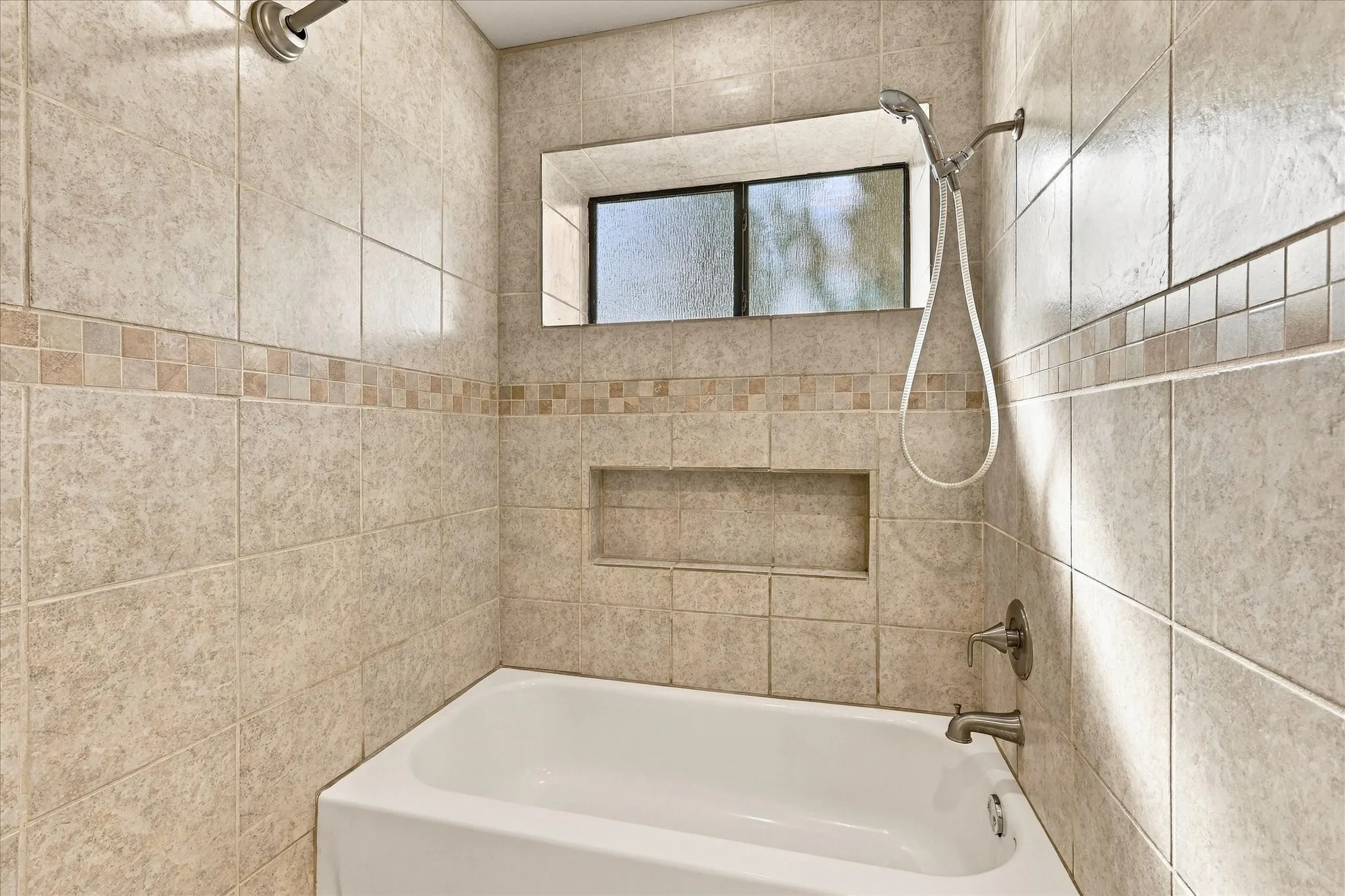 Bathtub and shower combination featuring full-height neutral ceramic tile surround, integrated tiled shower niche, decorative mosaic accent banding, and a privacy-glass window