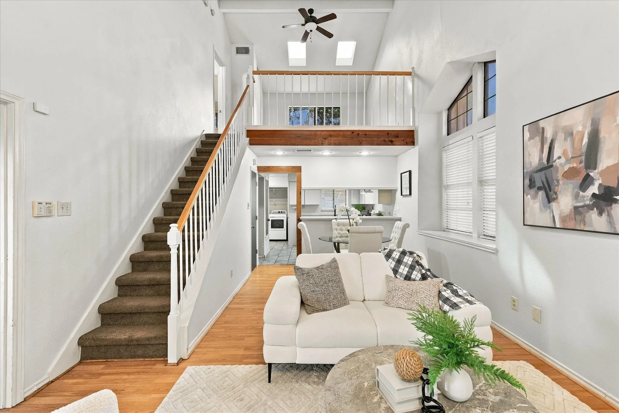 Vaulted ceilings with skylights, wood-finish flooring, a mezzanine level featuring a wooden railing, a staircase with white balusters, and oversized windows
