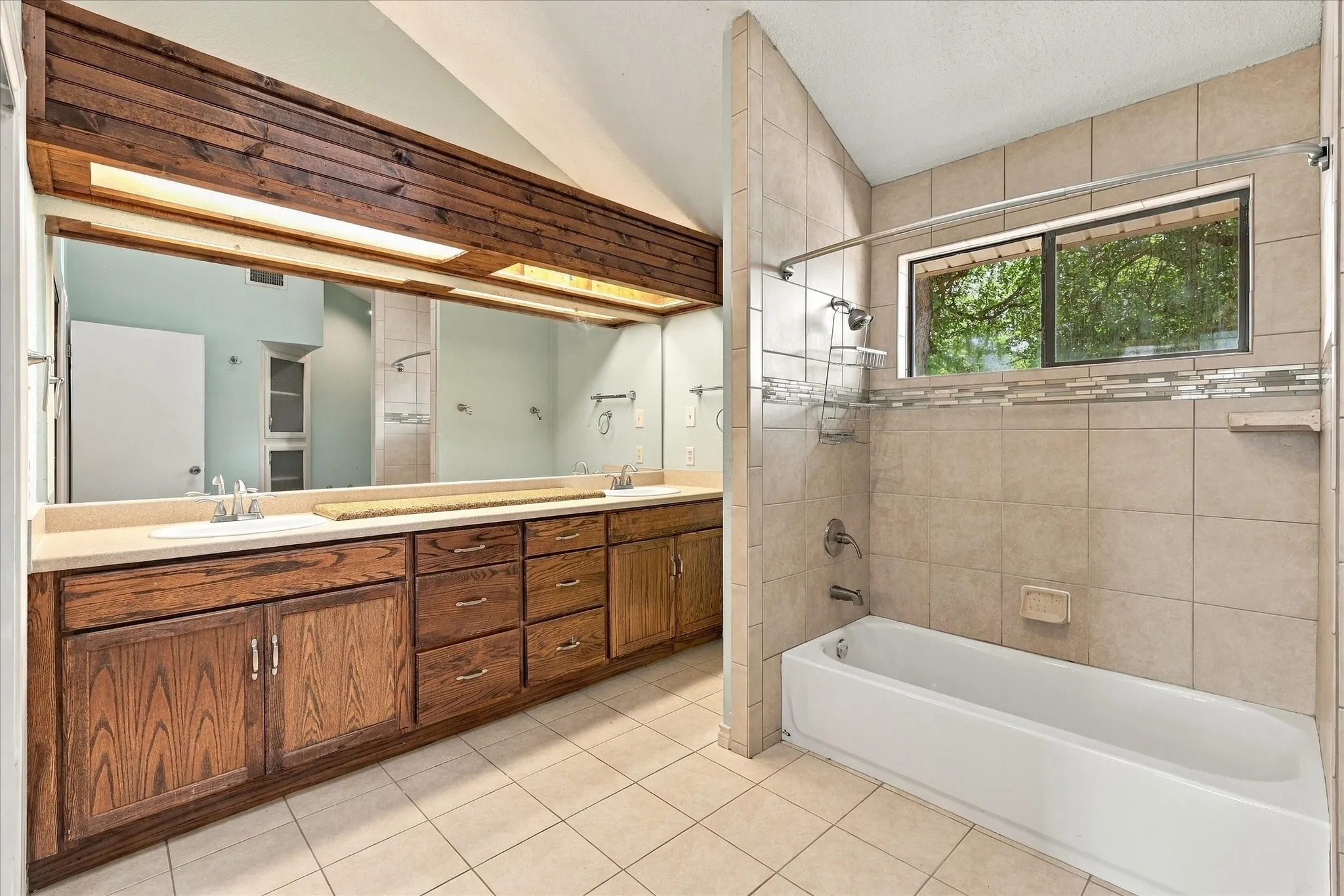 Full-width vanity with wooden cabinetry, dual integrated sinks, and expansive wall mirror featuring an integrated wooden valance with recessed lighting, paired with a tiled shower-tub combination