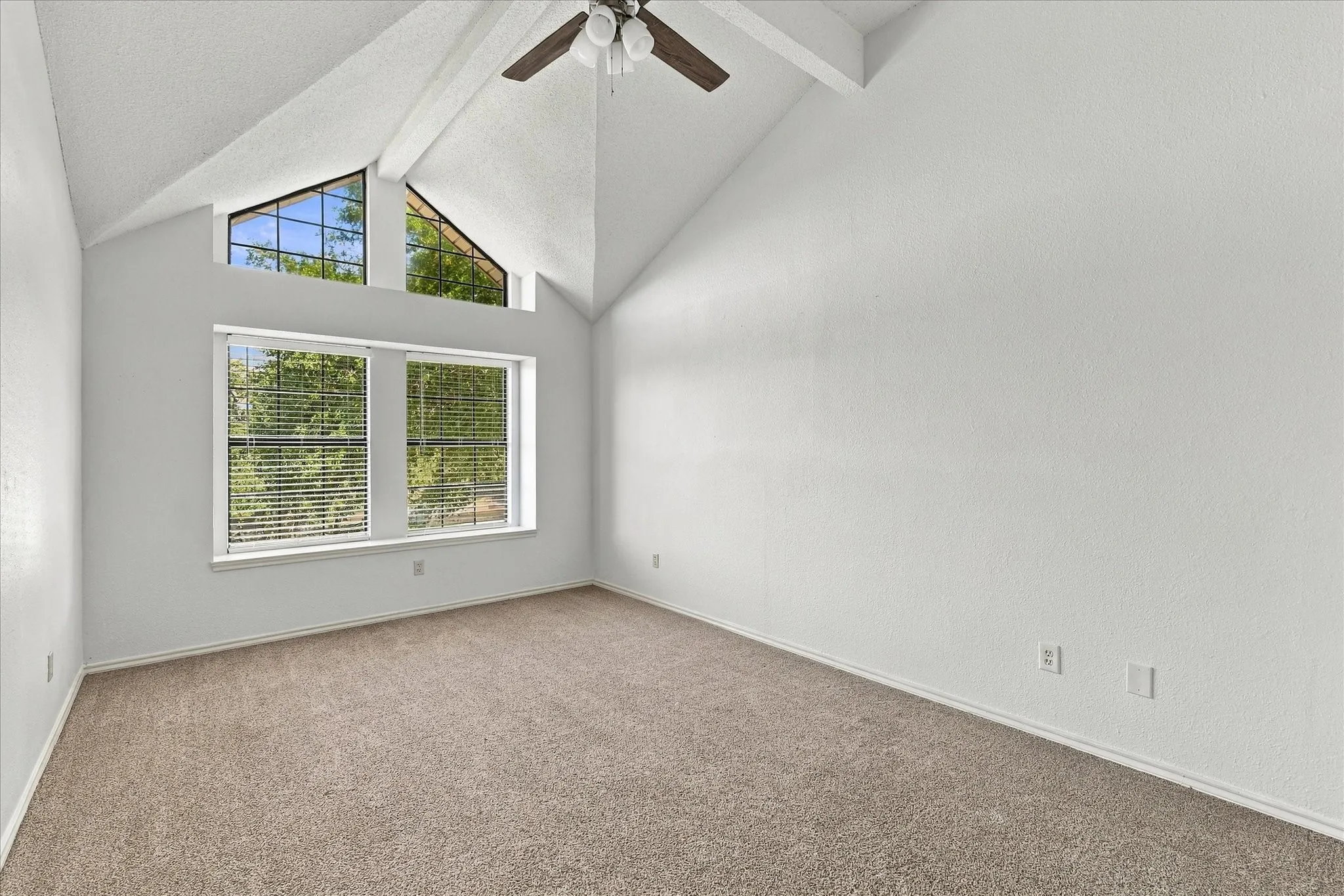 Vaulted ceiling featuring exposed beams, textured walls, neutral carpeting, multi-pane accent windows, and a ceiling fan