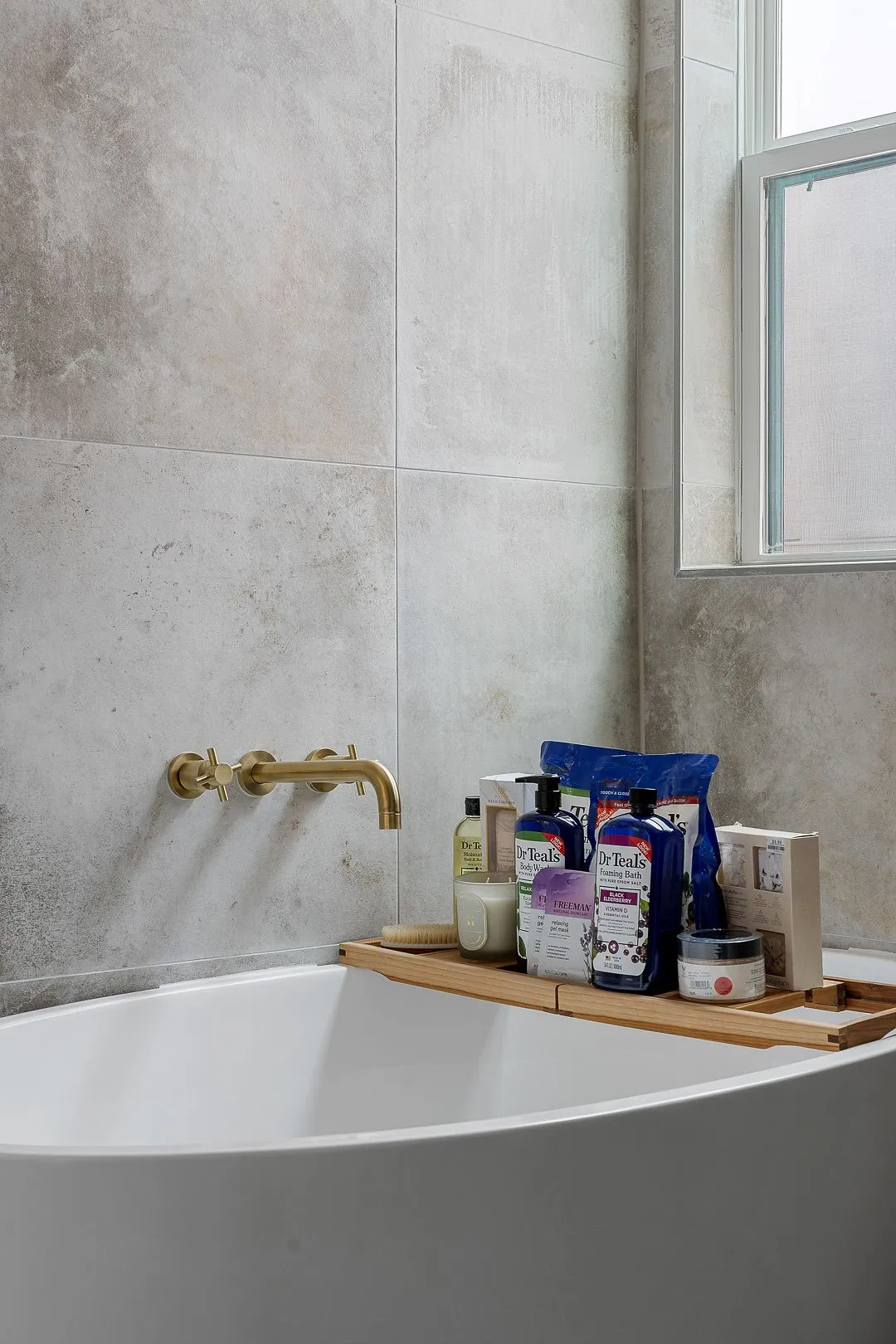 Freestanding soaking tub featuring large-format stone-finish wall tile, brass wall-mounted faucet hardware, and a window