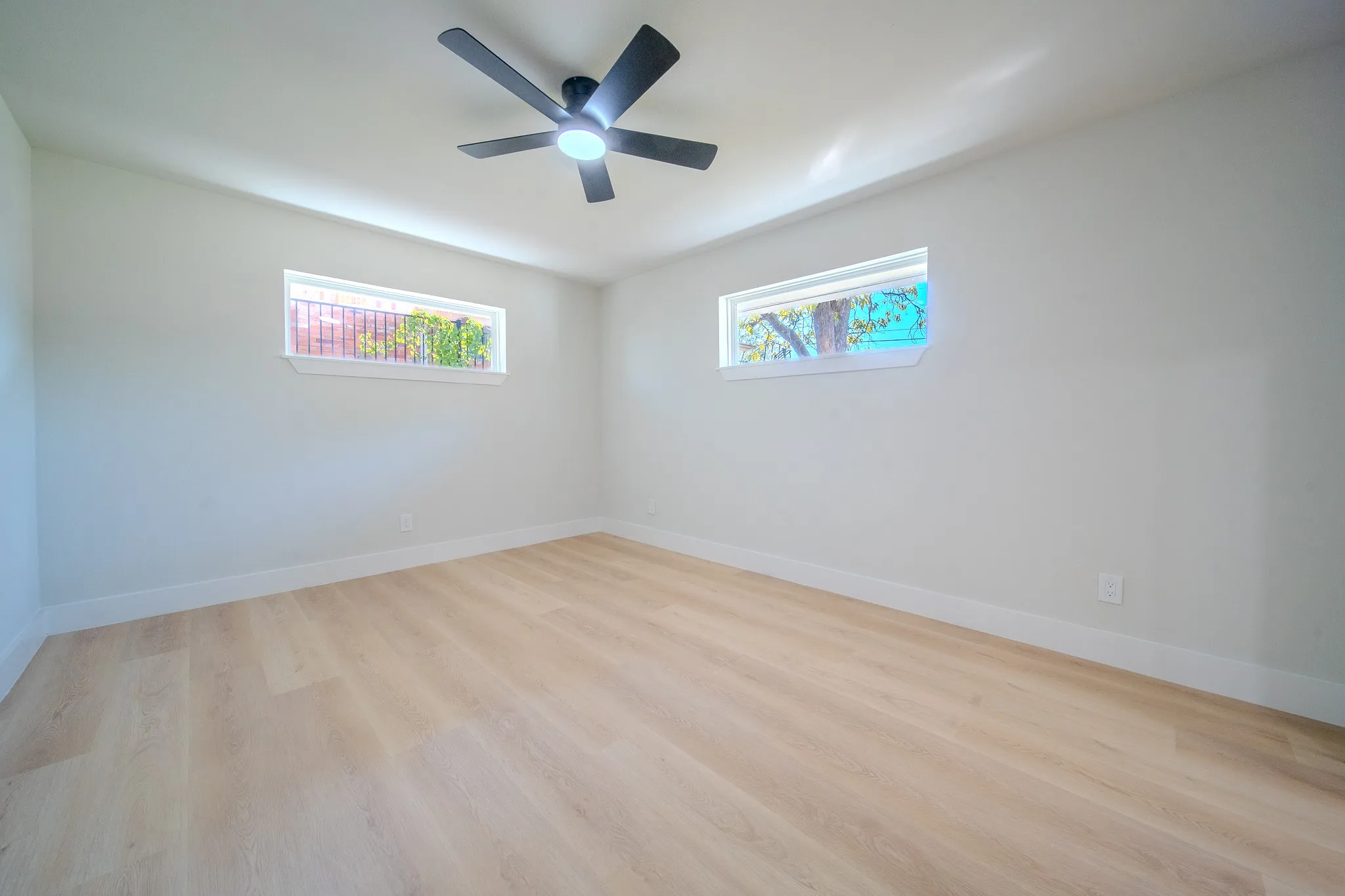 Neutral-toned interior room featuring wood-finish flooring, two horizontal rectangular windows, and a matte black ceiling fan with an integrated light fixture
