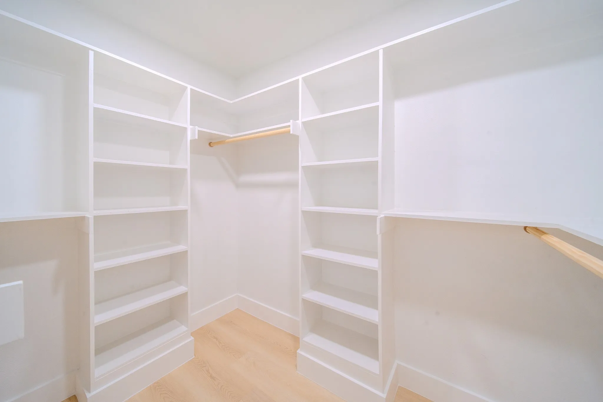 Custom walk-in closet featuring white built-in shelving, wooden hanging rods, and wood-finish flooring