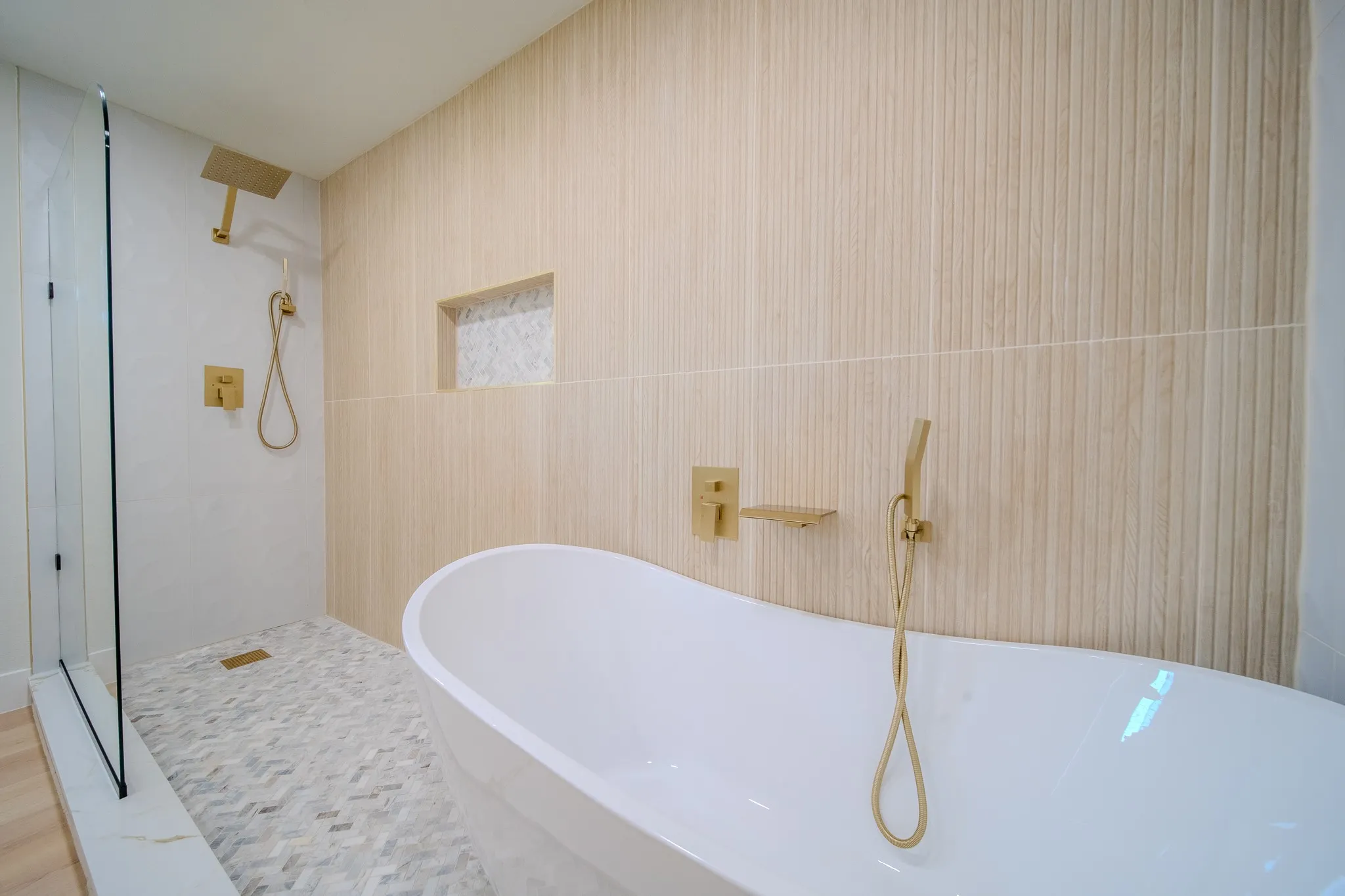 Freestanding soaking tub with gold-tone fixtures, walk-in shower with glass enclosure, vertically textured wall tiling, herringbone-patterned floor tile, and integrated wall niche