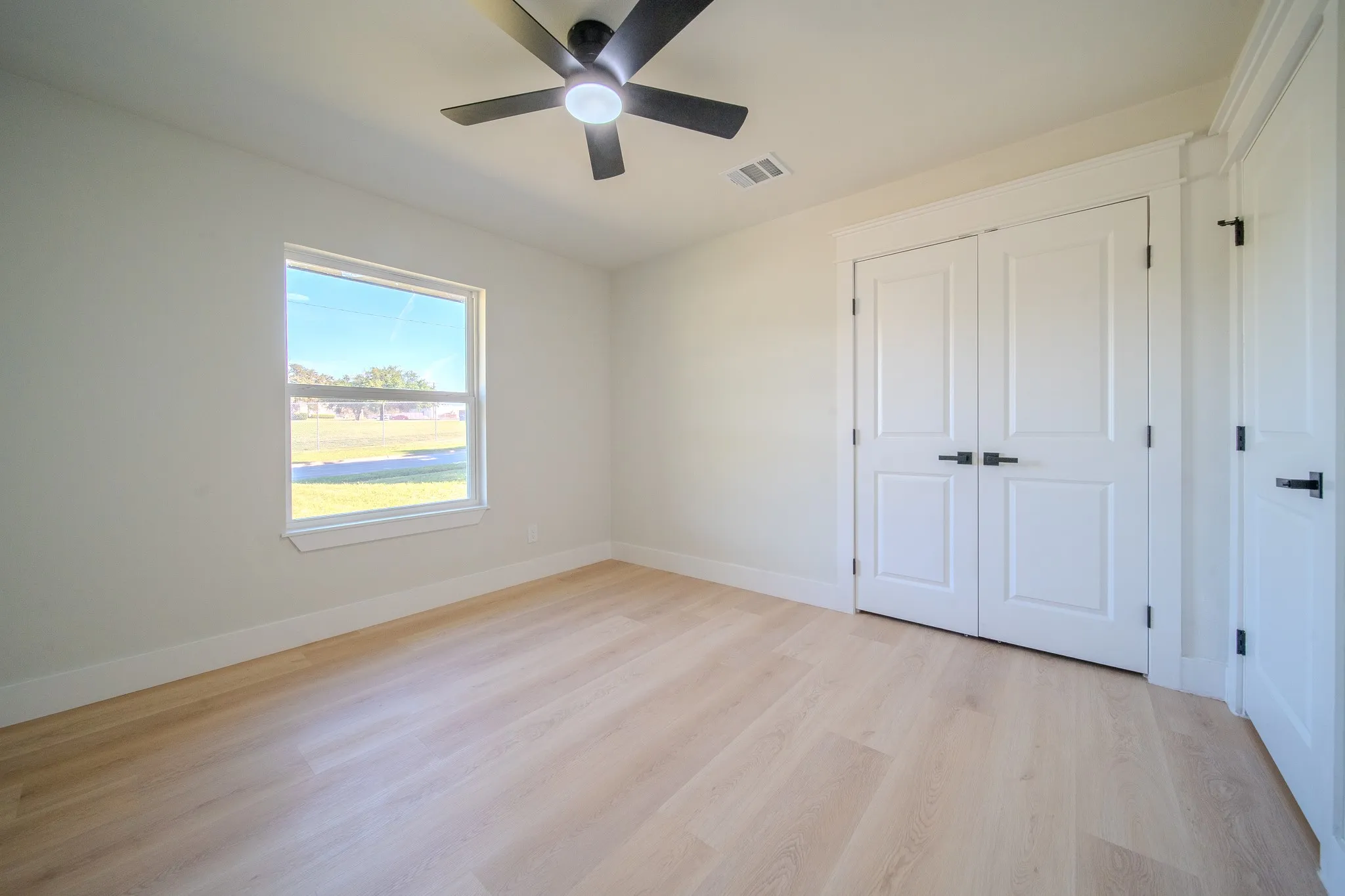 Neutral interior featuring light wood-finish flooring, double-panel closet doors with matte black hardware, and a ceiling fan with integrated lighting