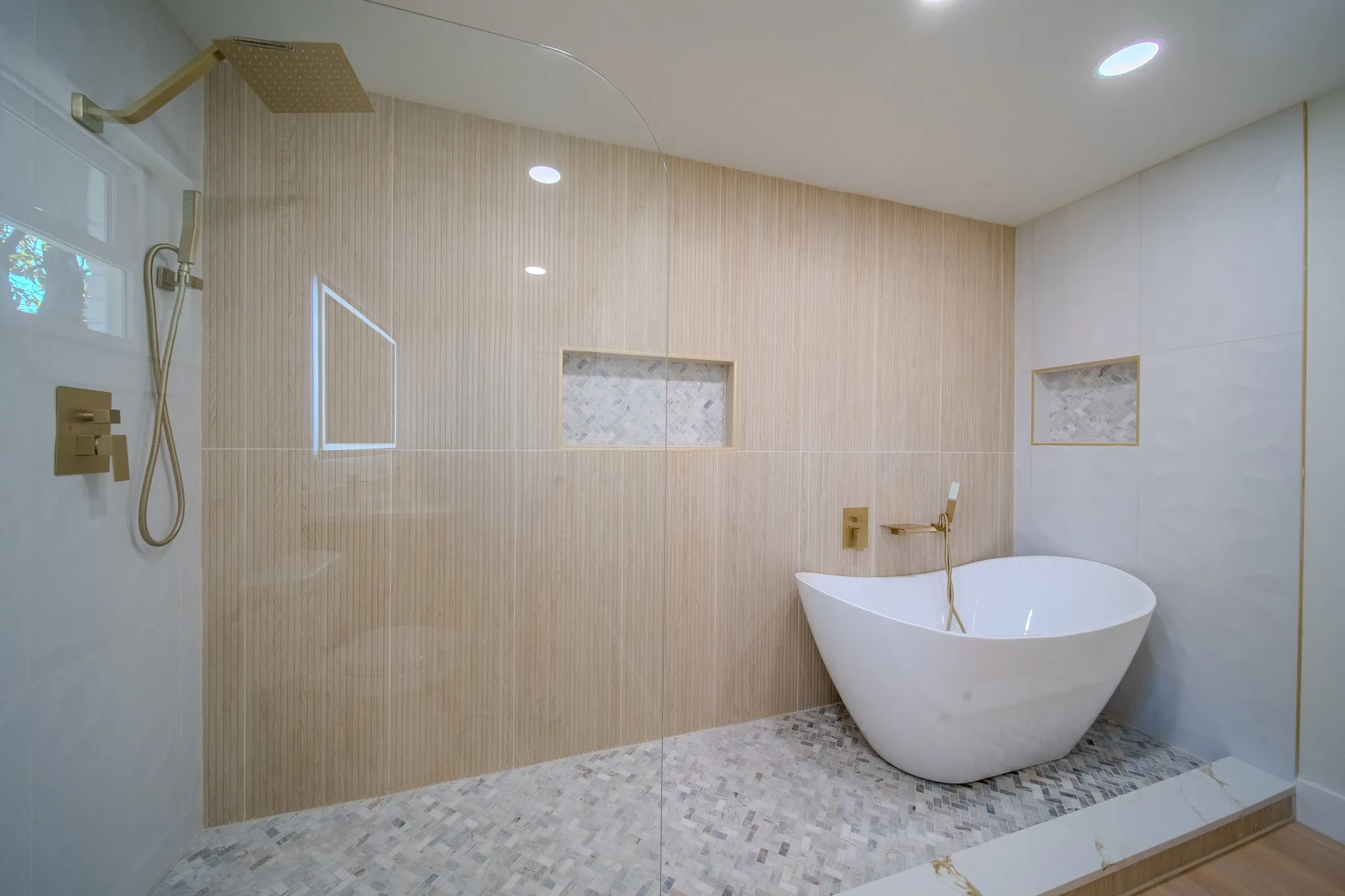 Freestanding soaking tub featuring gold-tone hardware, large-format textured wall tile, glass-enclosed shower area with rain-style showerhead, recessed wall niches, and intricate herringbone-patterned floor tile