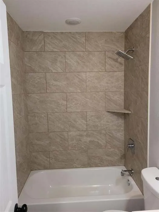 Bathtub and shower combination featuring large-format neutral-toned tile surround, built-in corner shelving, chrome rainfall showerhead, and matching chrome faucet fixture