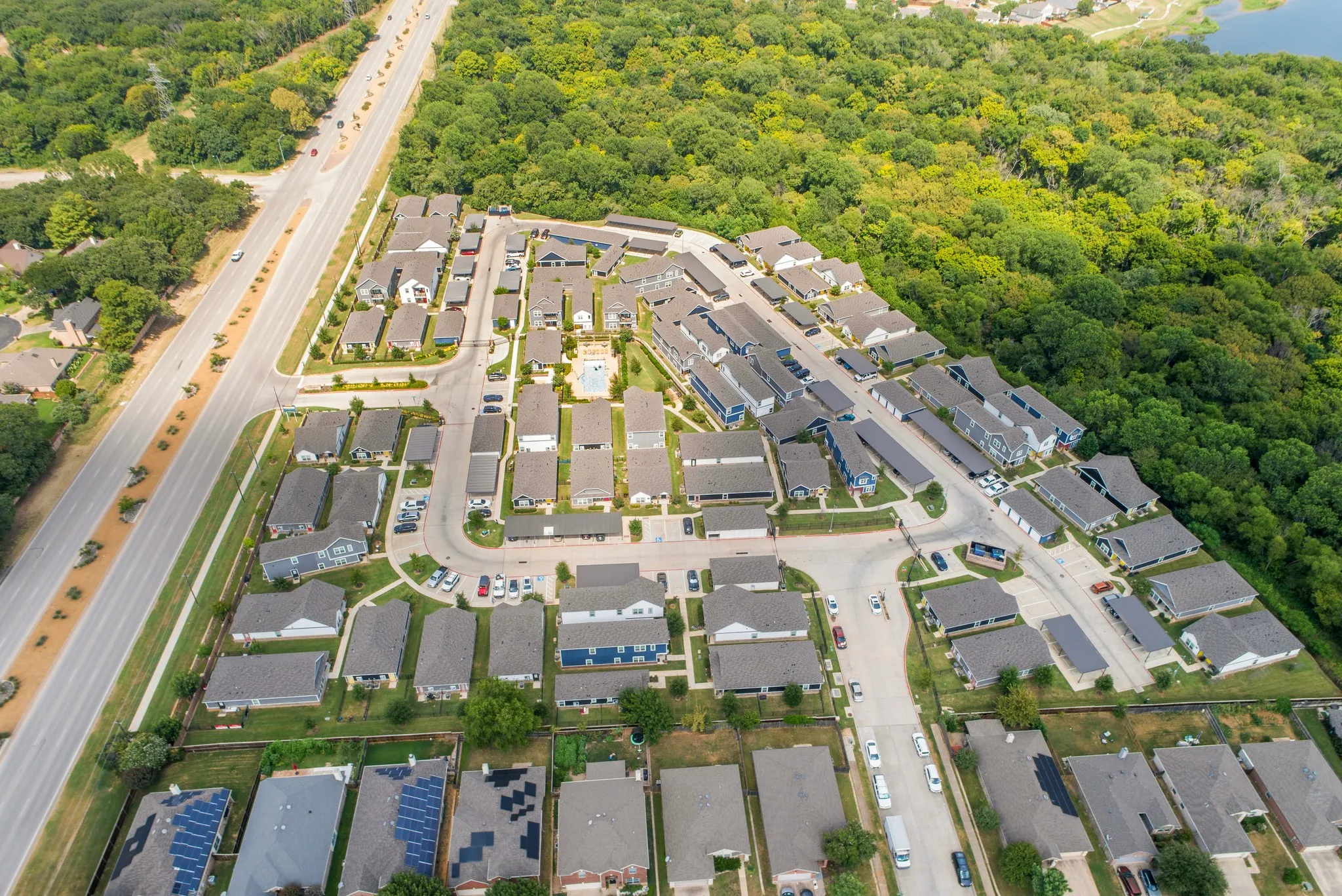 Aerial perspective of a multi-building residential community featuring a central swimming pool, covered parking structures, shingle-roofed architecture, and expansive surrounding woodland