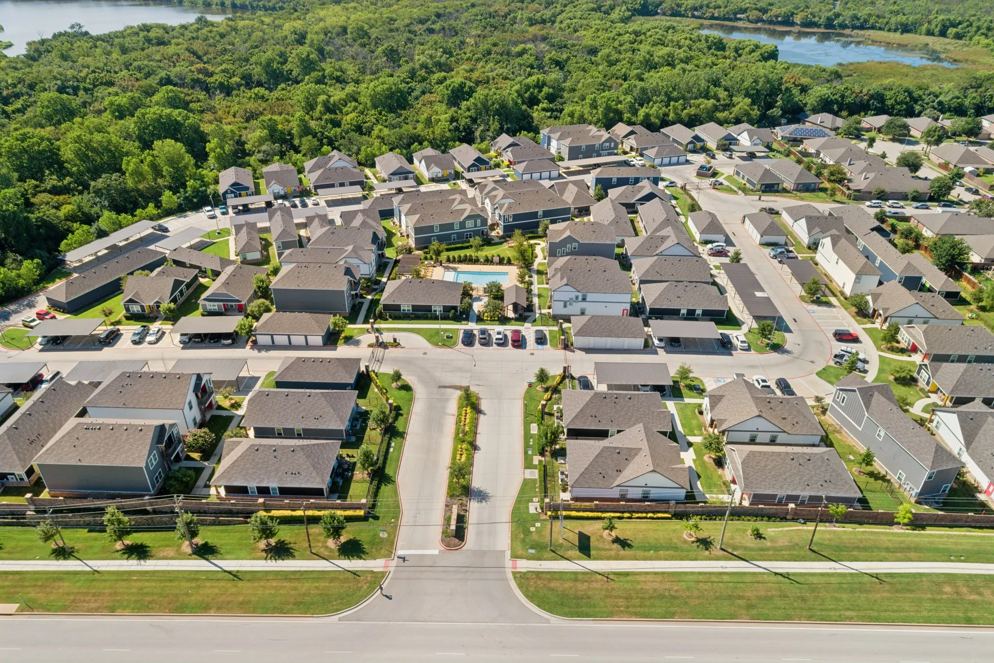 Aerial perspective of a multi-unit residential community featuring asphalt roadways, covered parking structures, a central swimming pool, and proximity to wooded green space and a pond