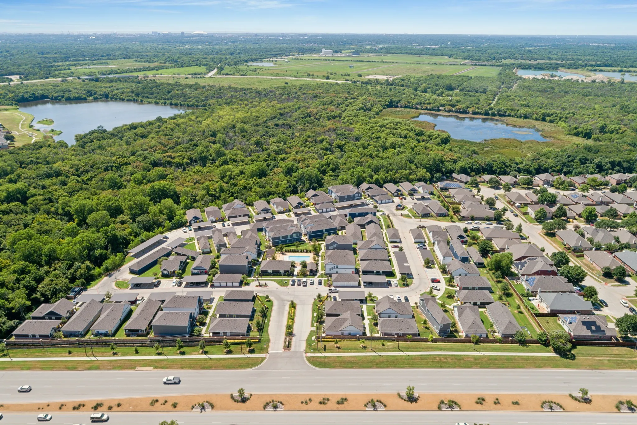 Aerial perspective of a multi-unit residential community featuring a central swimming pool, paved driveways, dense perimeter tree canopy, and nearby ponds