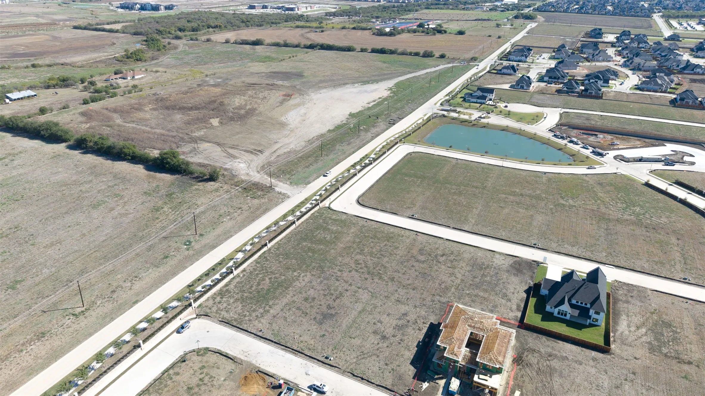 Aerial perspective of undeveloped land featuring paved road infrastructure, a retention pond, and perimeter fencing