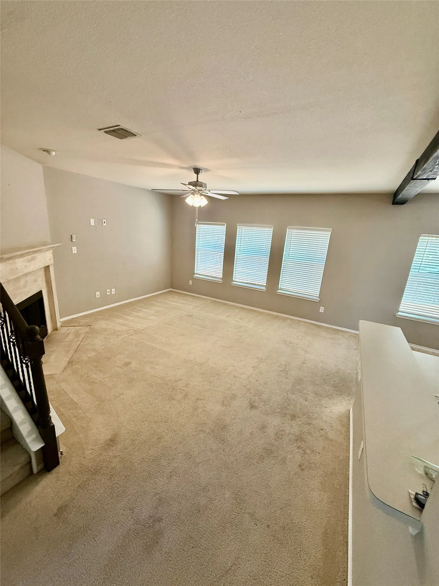 Living area featuring a fireplace with a light-toned surround, four windows with blinds, a ceiling fan with integrated lighting, and a dark-toned wood-finish banister