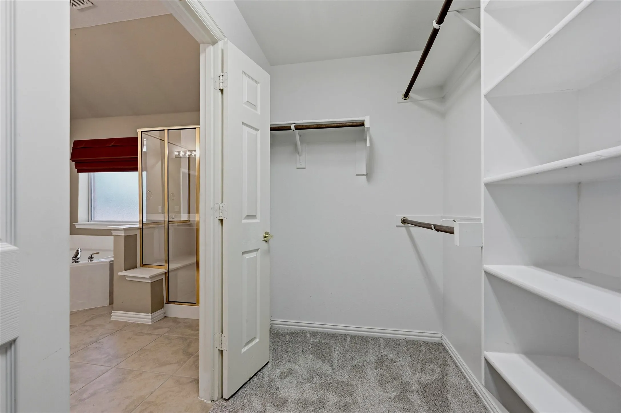 Walk-in closet featuring white built-in shelving and dual hanging rods, with tiled flooring and a glass-enclosed shower visible beyond the doorway
