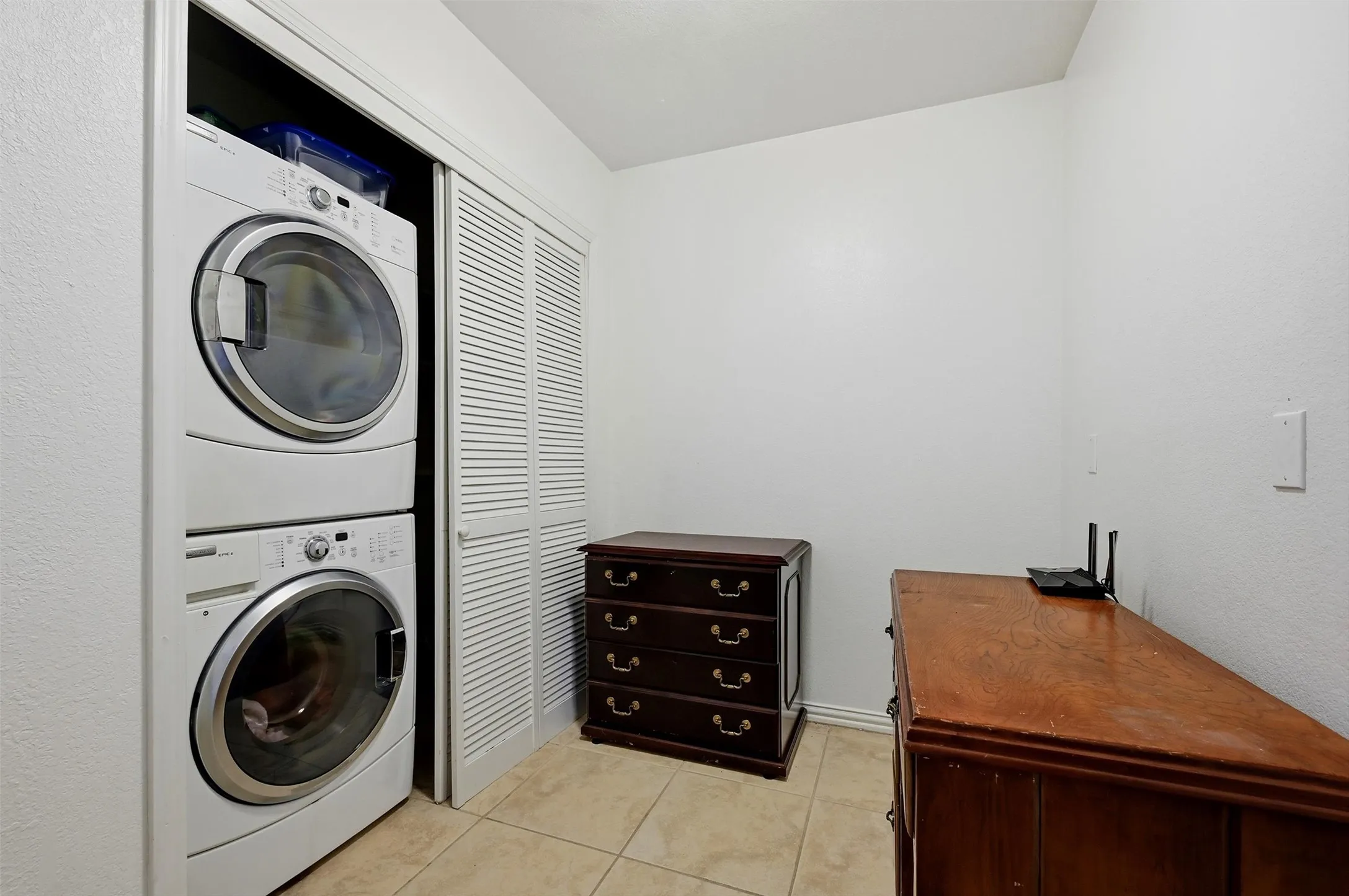 Laundry area featuring stacked front-load washer and dryer, white louvered bifold closet doors, and neutral square tile flooring