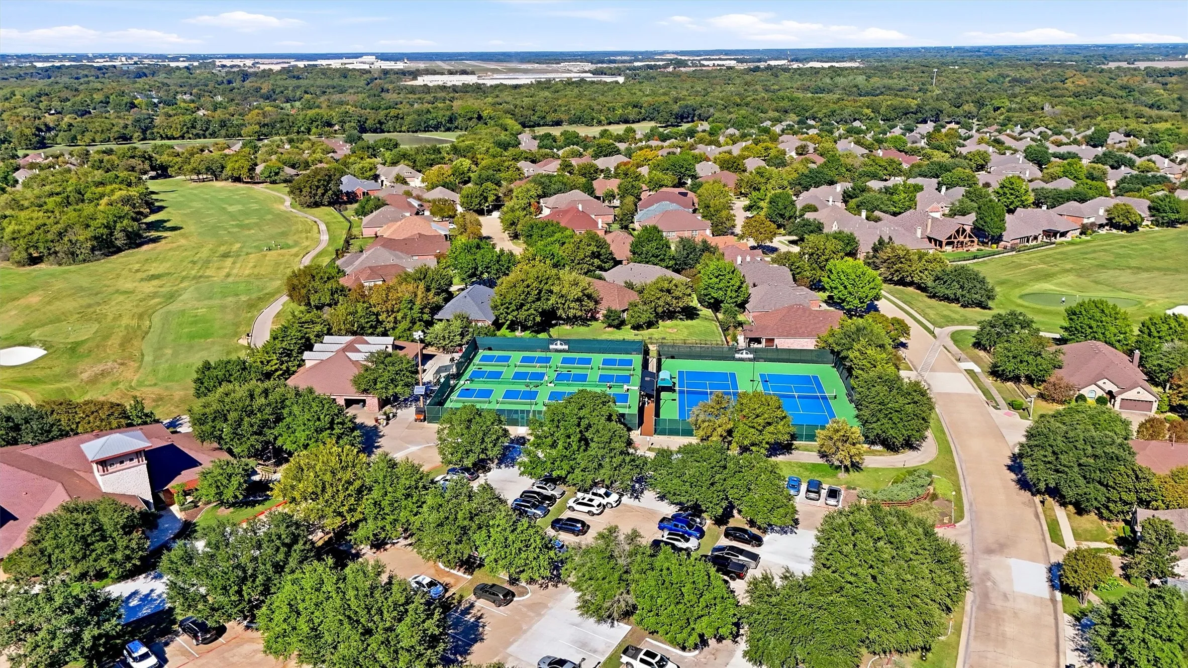 Aerial perspective of tennis courts, paved parking area, and surrounding green space with mature trees