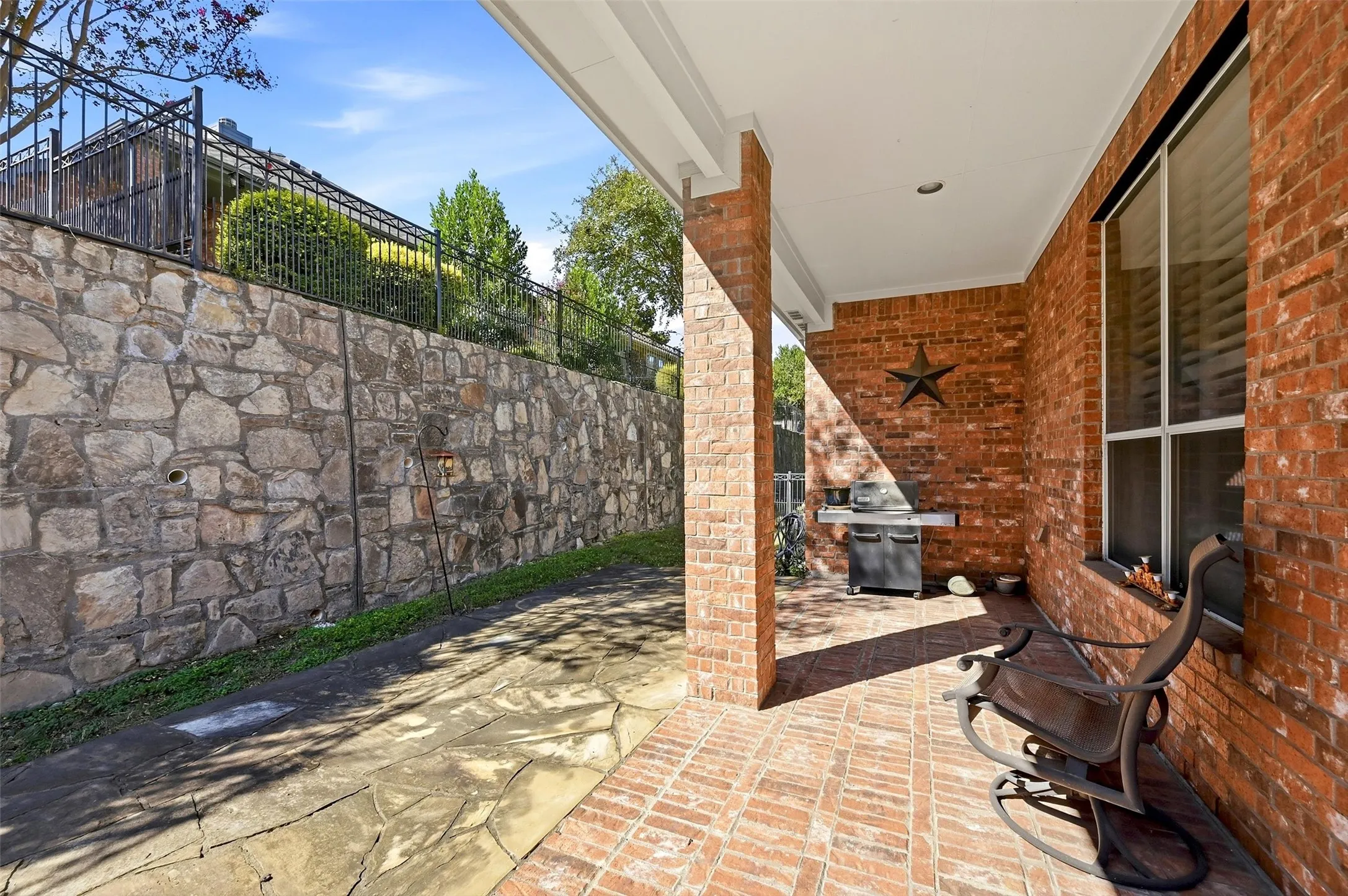Covered patio featuring red brick flooring and a masonry column, complemented by a natural stone retaining wall and a black metal perimeter fence