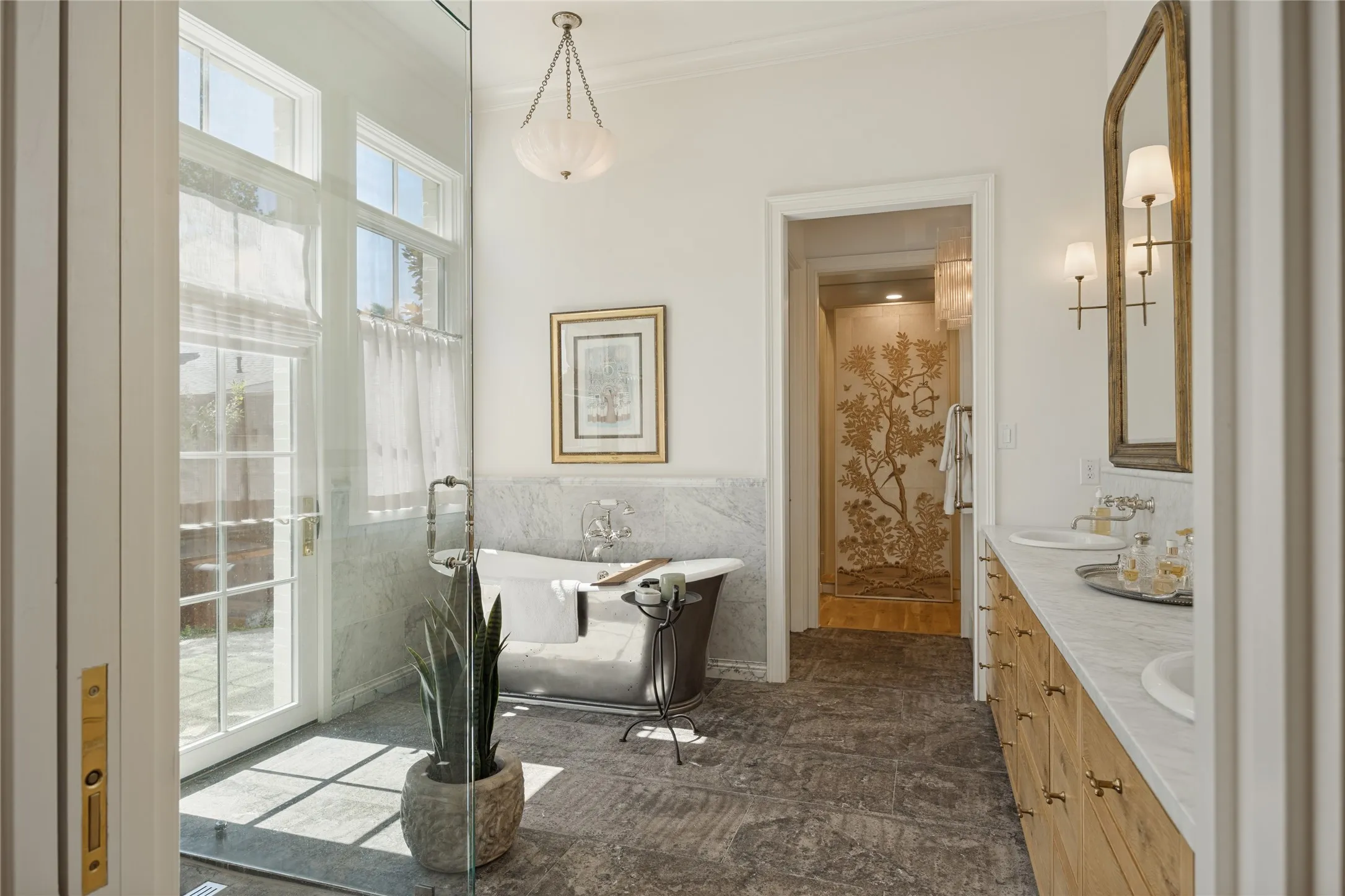 Freestanding soaking tub with stone surround, dual-sink wooden vanity with stone countertop, natural stone tile flooring, glass-enclosed shower, and multi-light pendant chandelier