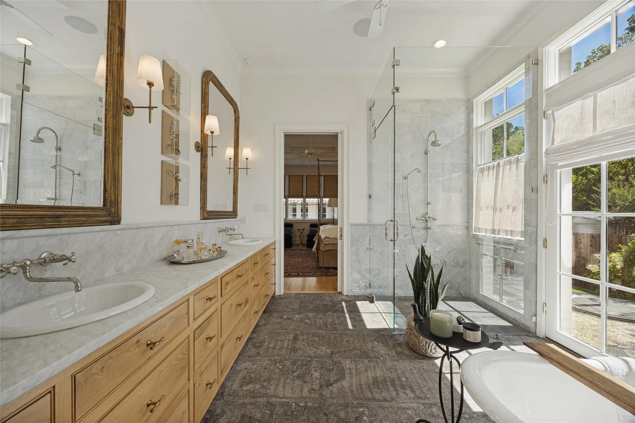 Custom wood-finish vanity with dual integrated sinks and stone countertops, stone flooring, frameless glass-enclosed shower with stone surround, and freestanding soaking tub