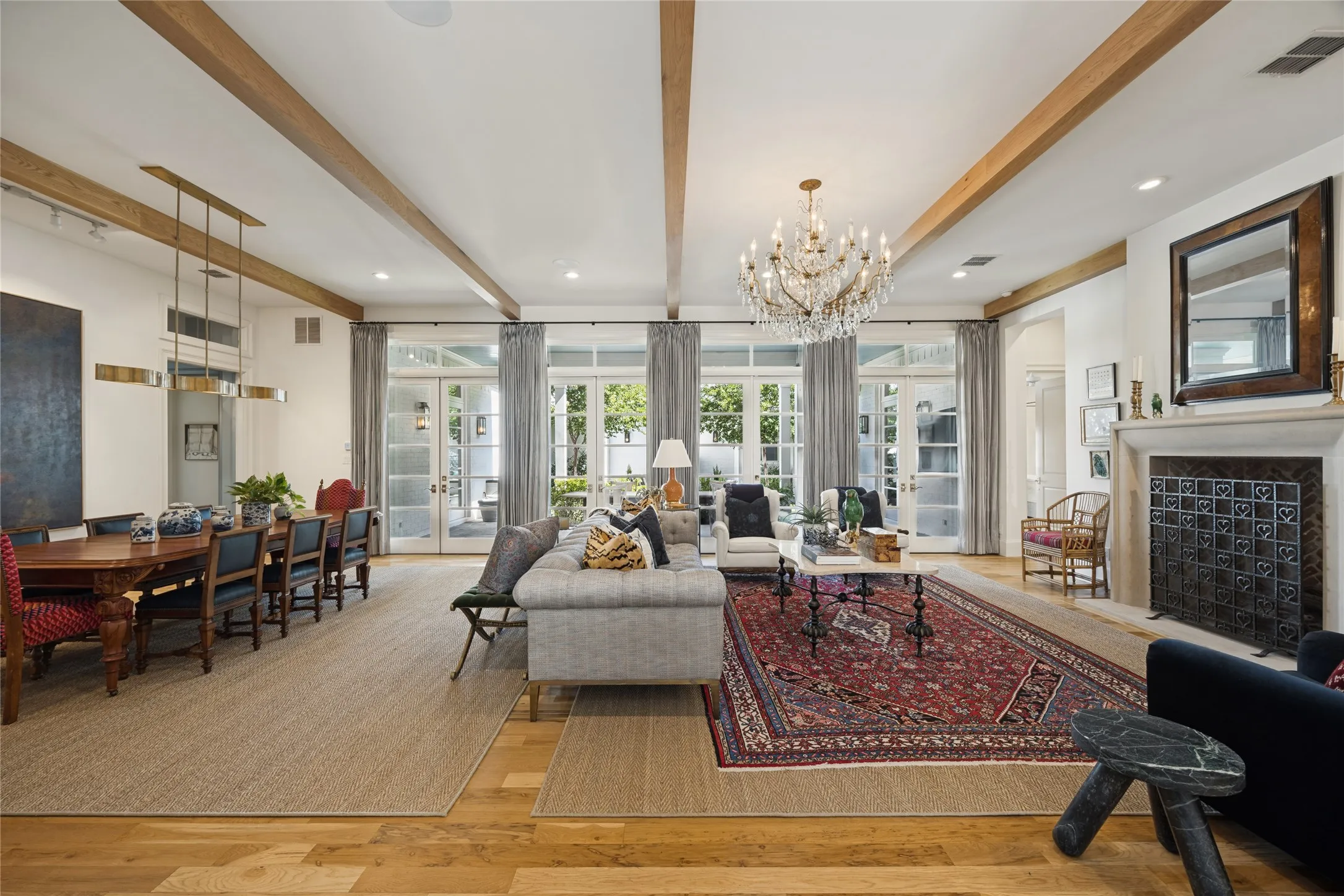 Open-concept space featuring wood-finish flooring, exposed ceiling beams, a masonry fireplace with an ornate metal screen, multi-pane glass-panel doors, and a crystal chandelier