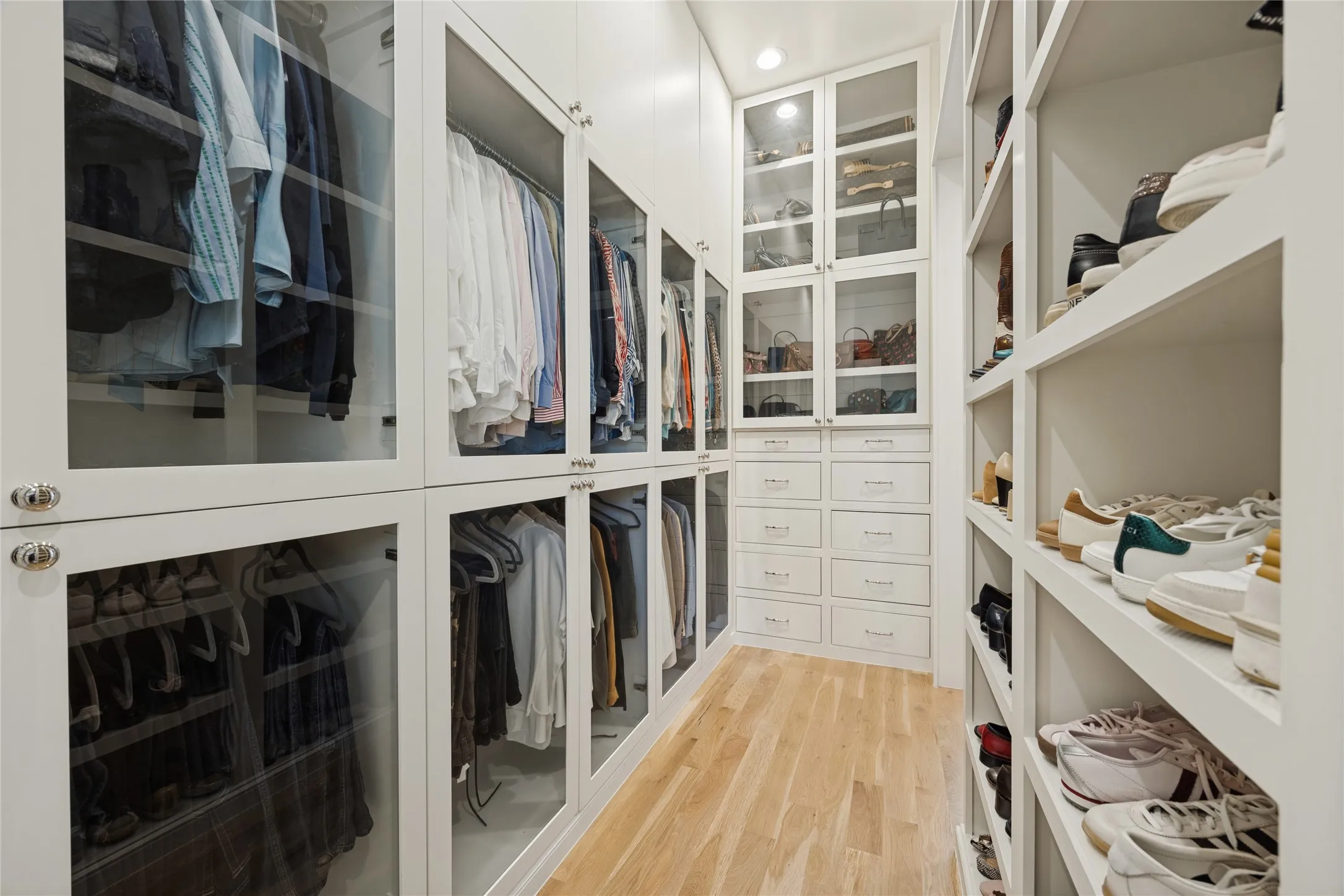Custom walk-in closet featuring white built-in cabinetry with glass-front doors, integrated drawers, open shelving, and wood-finish flooring