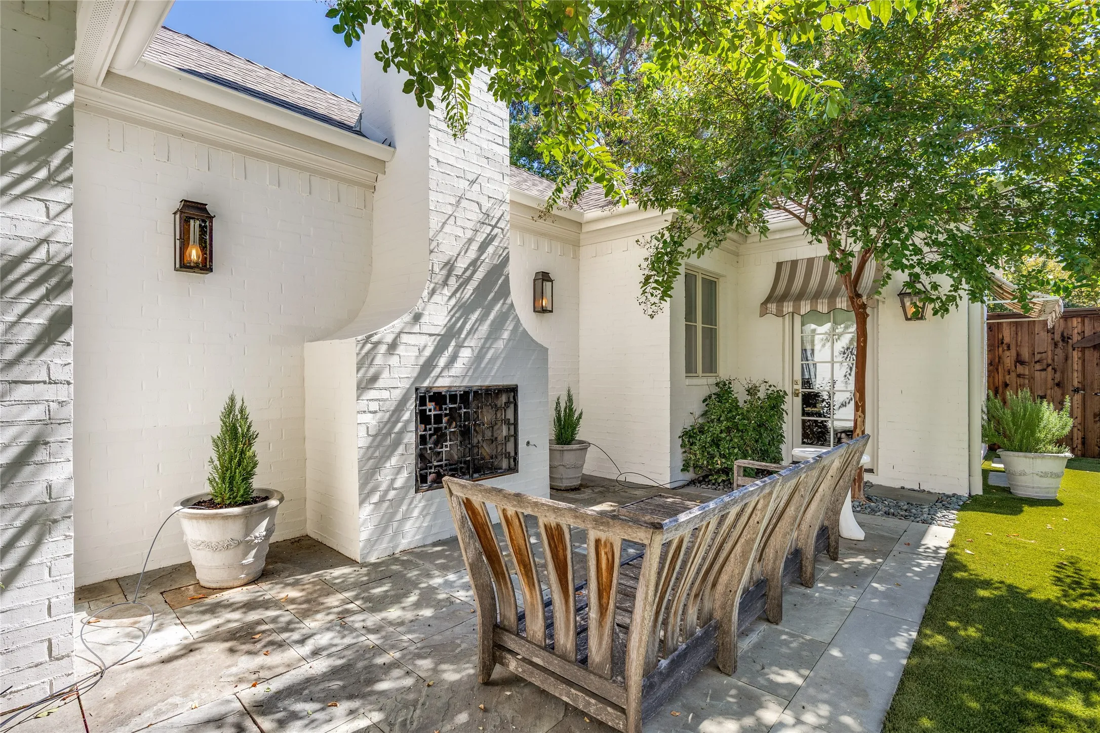 Outdoor patio featuring a white brick masonry fireplace, natural stone flooring, wall-mounted exterior lanterns, and a striped fabric awning over the entryway