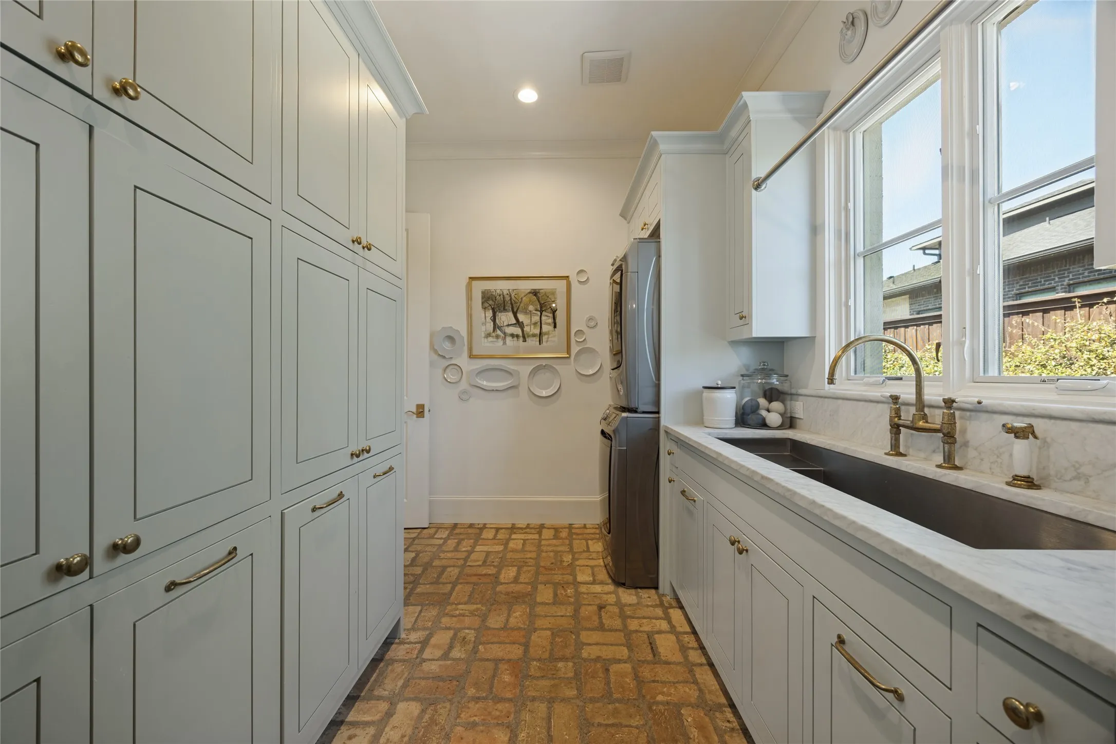 Laundry room featuring full-height light-colored cabinetry, brick flooring, a farmhouse sink with brass-finish hardware, stone countertops, and a window