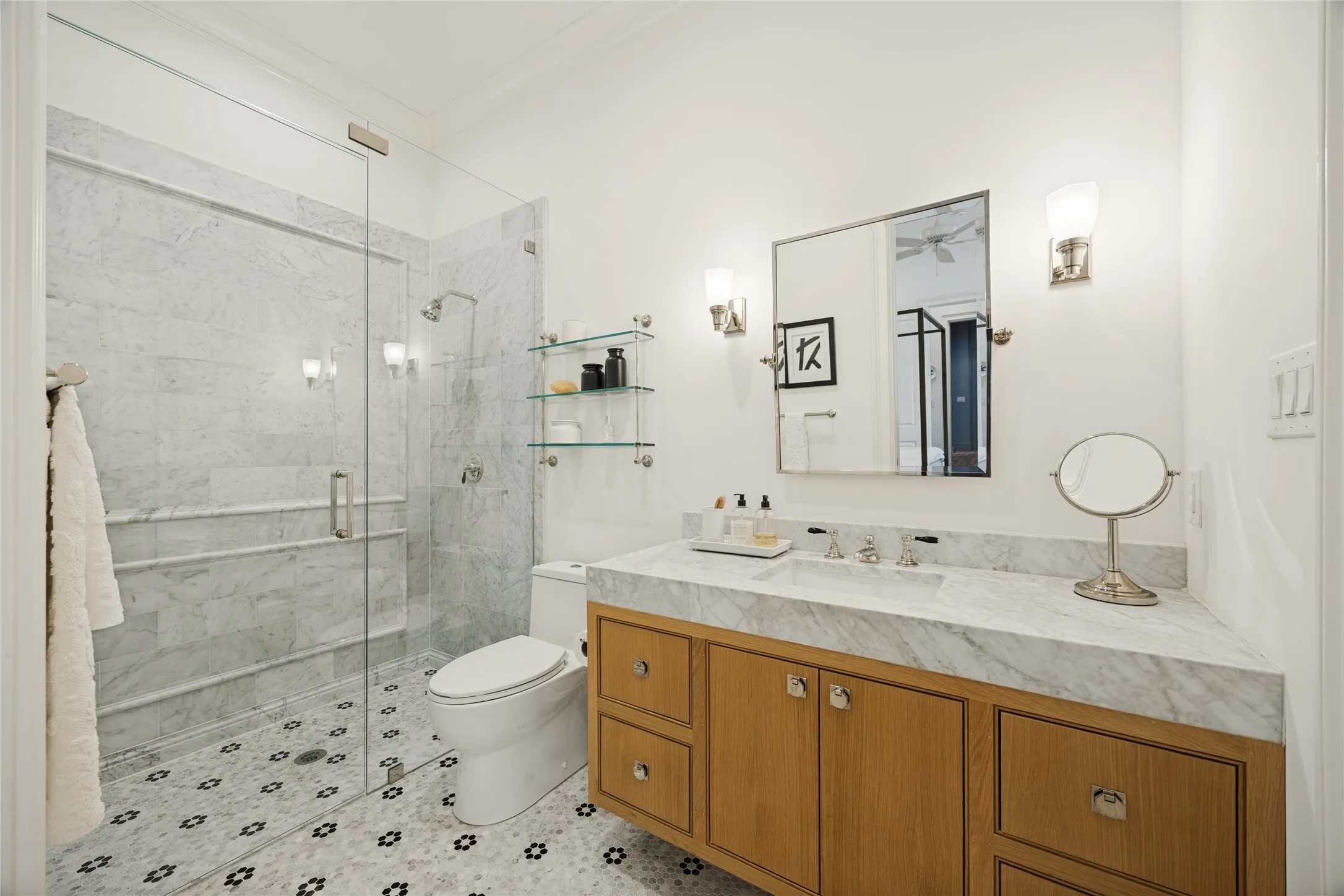 Bathroom featuring a walk-in shower with stone surround, stone-topped vanity with natural wood-finish cabinetry, patterned mosaic tile flooring, and wall-mounted light fixtures