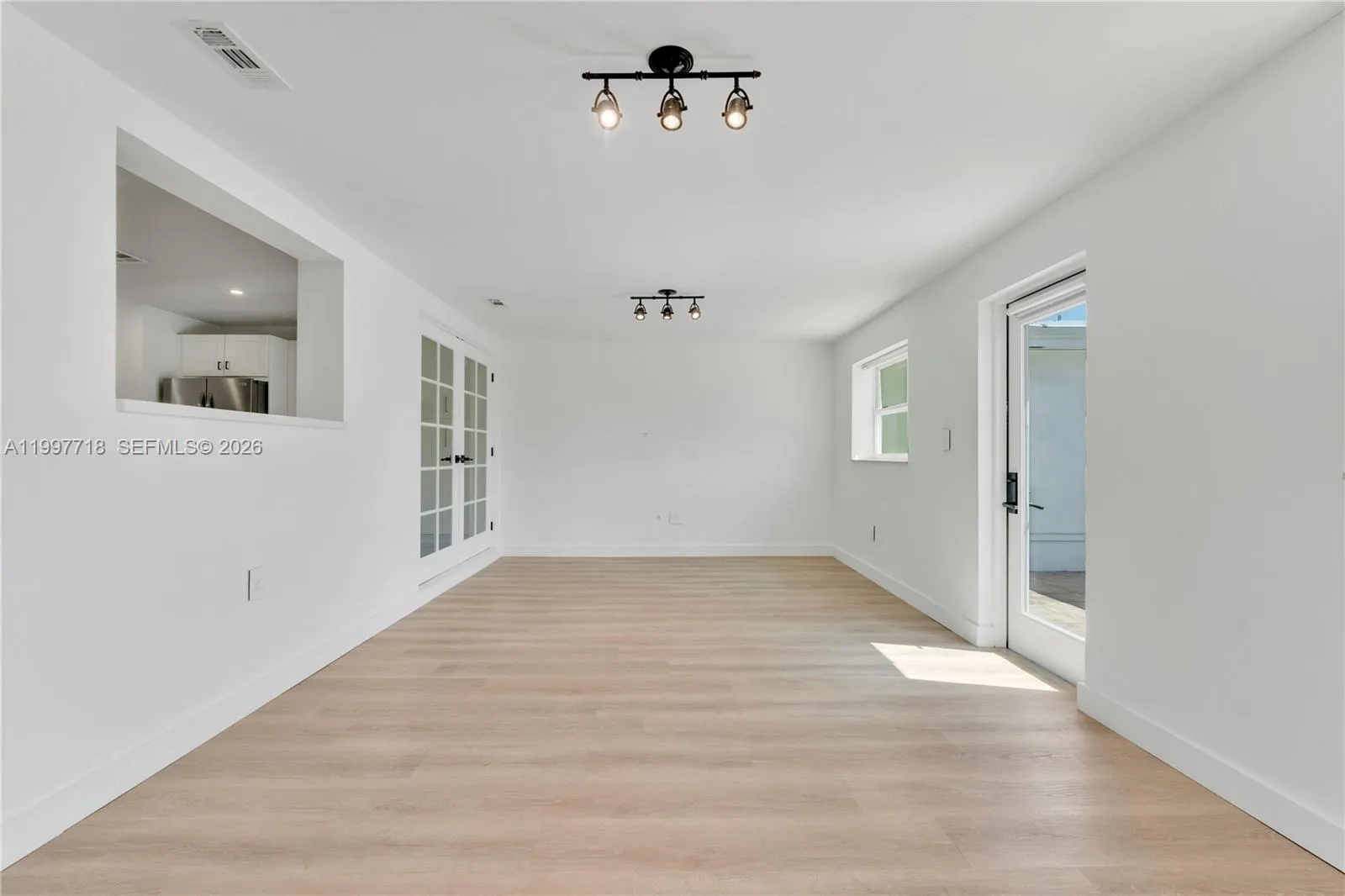 A long, open living space with light hardwood-style flooring, track lighting, and a kitchen pass-through window on the left. Features French glass doors leading to another room, plus a side door opening to the outdoor area.