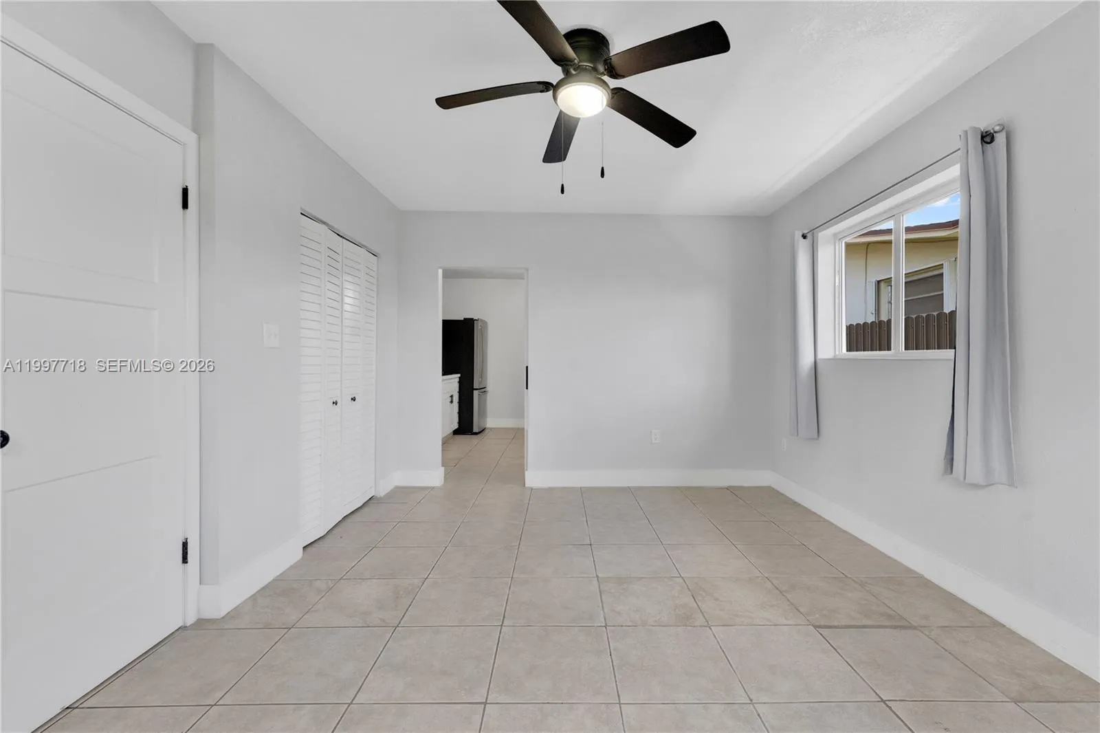A spacious, empty bedroom with light gray walls, large tile flooring, a ceiling fan with light, two closets (one louvered, one solid door), a side window with curtains, and an open doorway leading to the kitchen area.