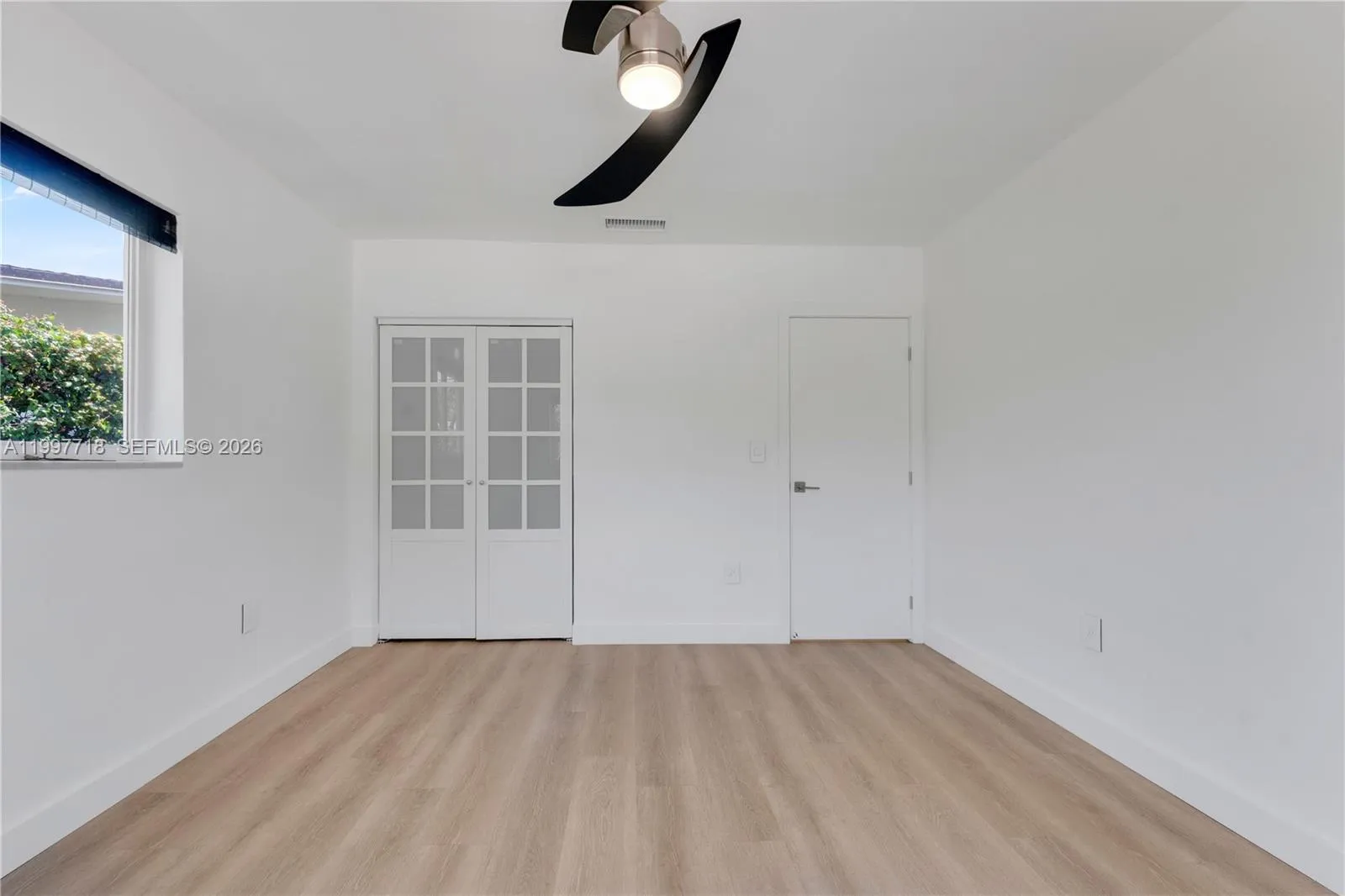 A bright, empty bedroom with light hardwood-style flooring, white walls, a modern two-blade ceiling fan, and two windows with roller shades offering views of the neighborhood outside.