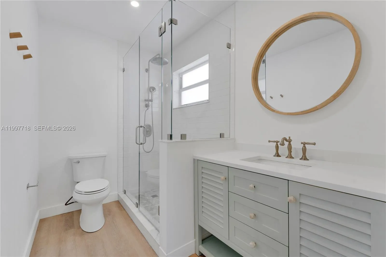 A elegant bathroom featuring a frameless glass walk-in shower with rain head, a sage green louvered vanity with white countertop and brushed gold faucet, a round wood-framed mirror, and light hardwood flooring.