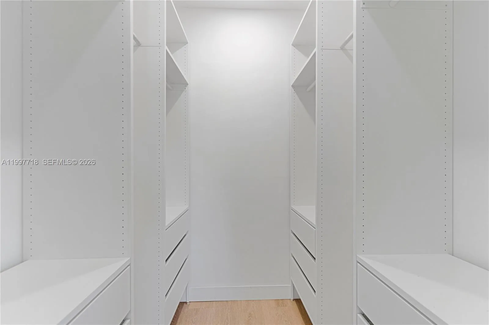 A clean, all-white walk-in closet with built-in shelving, hanging rods, and drawers on both sides, offering ample organized storage space. Light wood flooring runs along the floor.